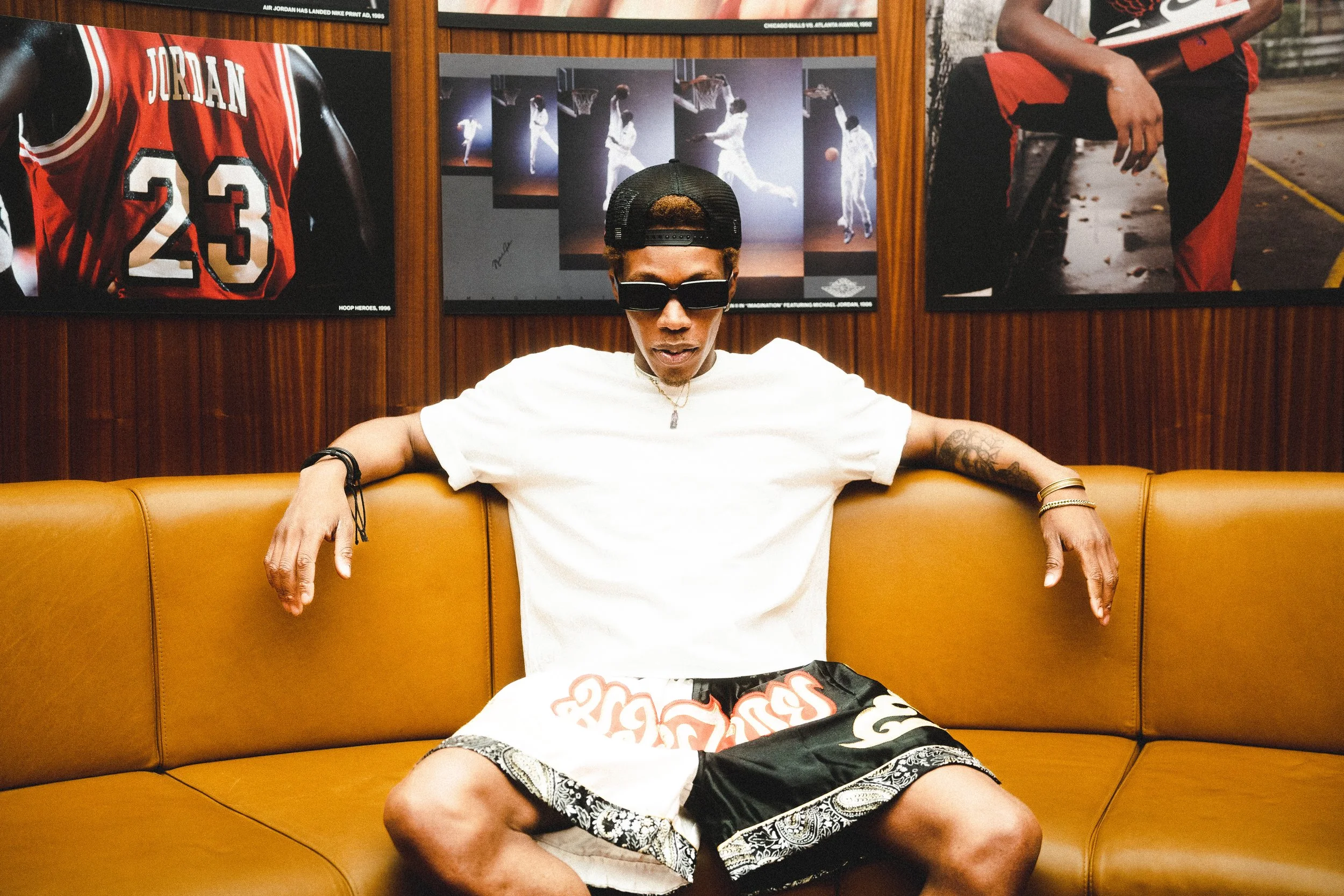 A young man wearing a black cap, dark sunglasses, a white T-shirt, and patterned shorts sits on a mustard-colored leather couch with his arms stretched out. He has tattoos on his arms and is adorned with jewelry. Behind him are framed photographs of 