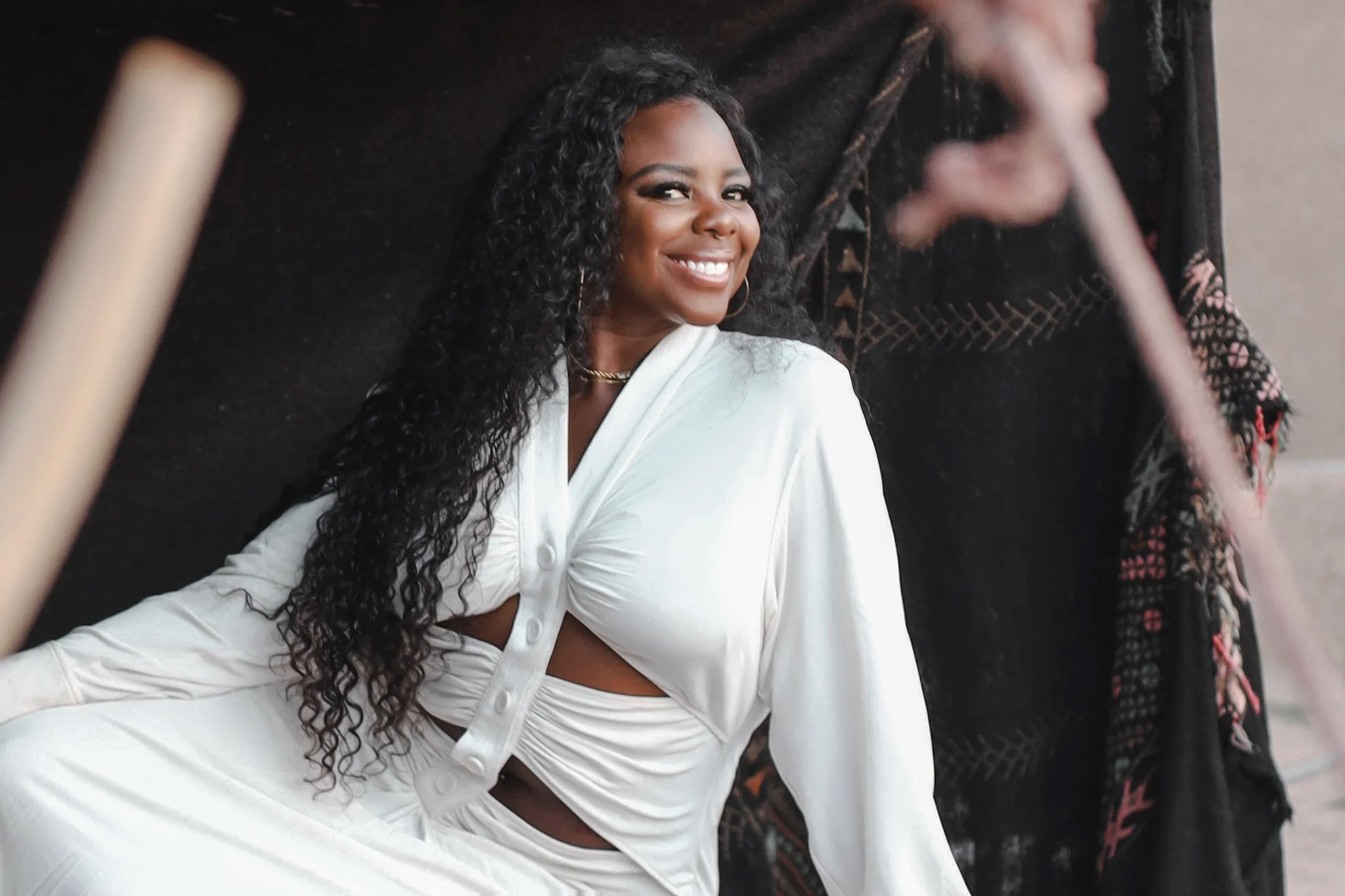 A woman with long curly black hair, smiling, wearing a white outfit, sitting next to another person who is partially visible and blurred in the background.