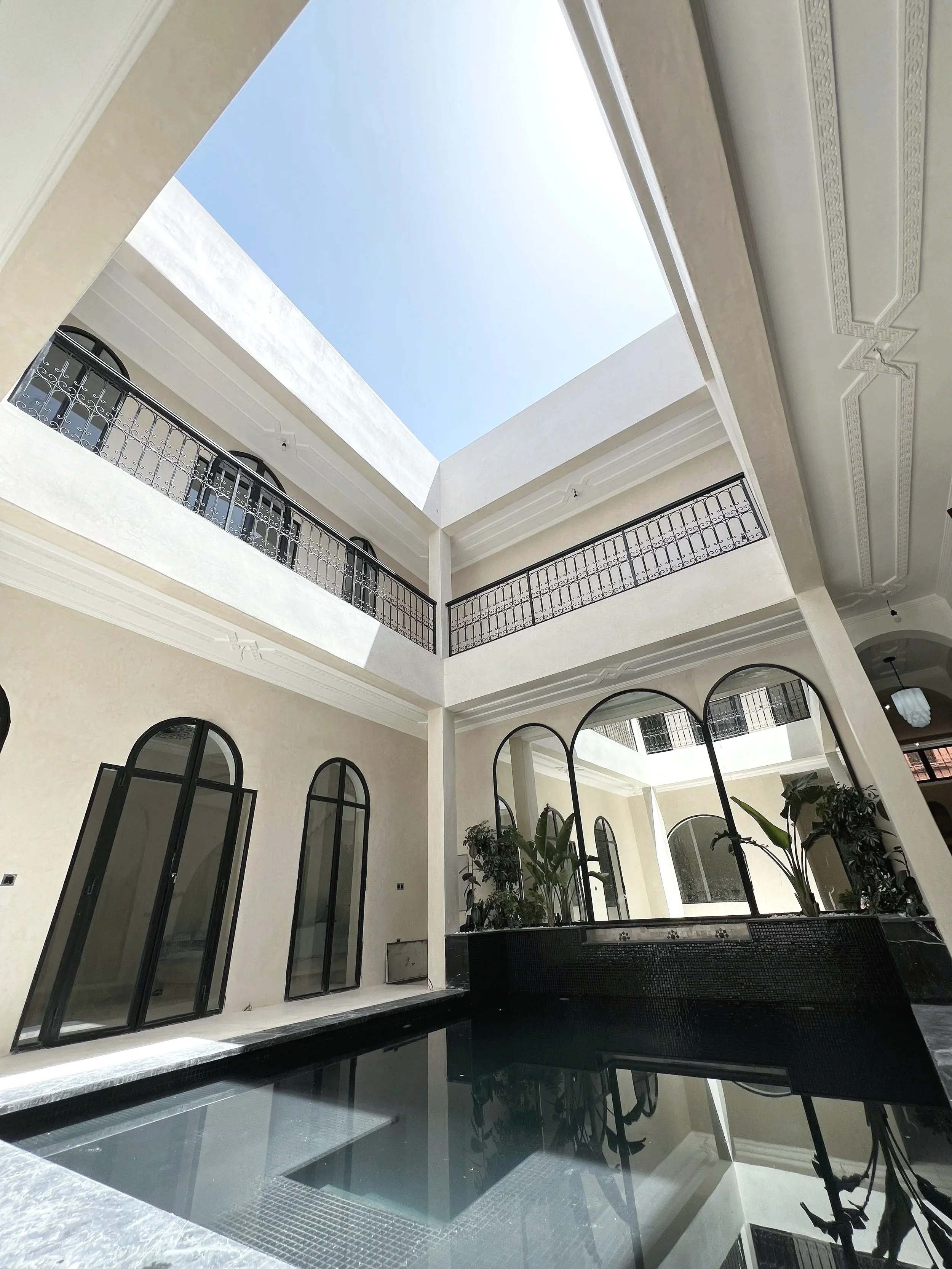 Interior view of a modern multi-story building with a rectangular open ceiling showing the sky, black balconies with decorative railings, large arched windows, indoor plants, and a black sectional sofa.
