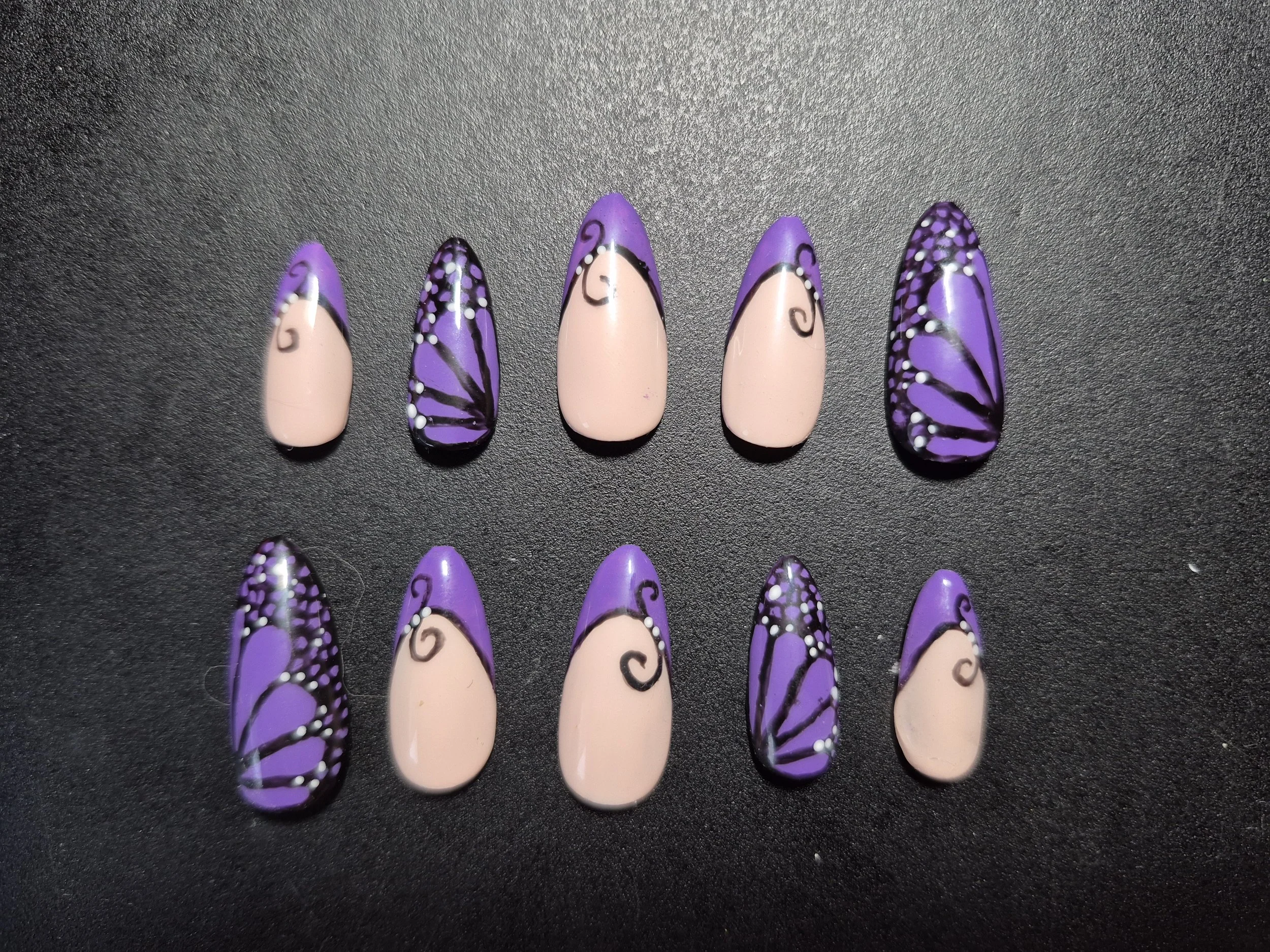 A row of artificial nails painted with purple and beige, featuring intricate black and white swirl and dot designs.