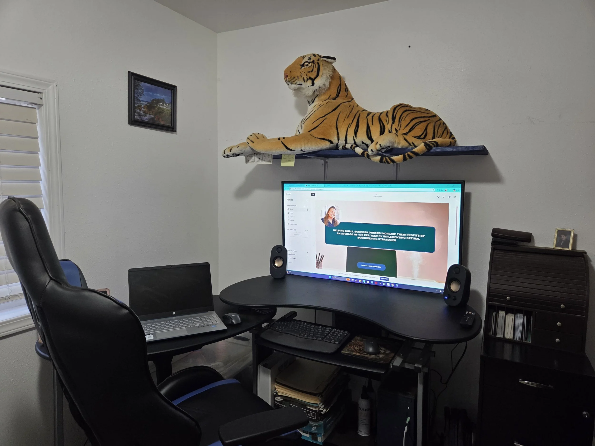 Home office with a computer desk, a large monitor displaying a website, a laptop, and a black office chair. A stuffed tiger is on a wall shelf above the monitor, and a small framed picture is on a dark wooden cabinet.