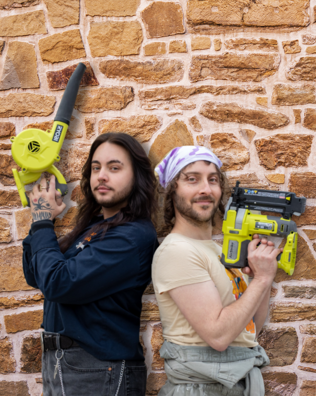 Two trans queer handy workers standing back to back with their arms crossed Charlies Angel style holding different tools , leaning against a brick wall.