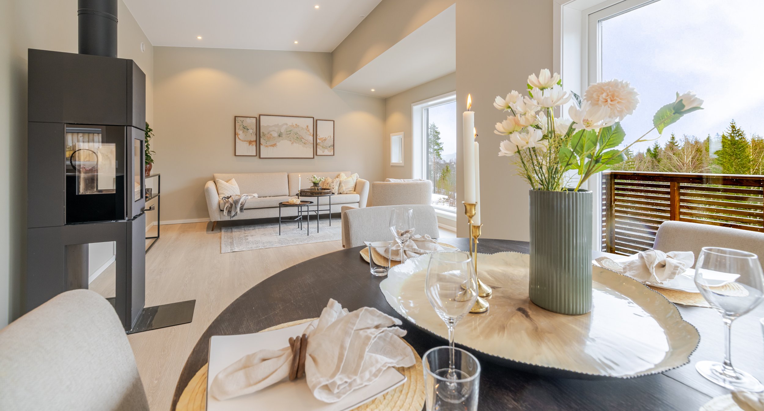 Et moderne stuebord med en blomstervase med hvite blomster, lysende stearinlys og spiseutstyr, satt opp foran store vinduer med utsikt til en terrasse og grønt landskap.