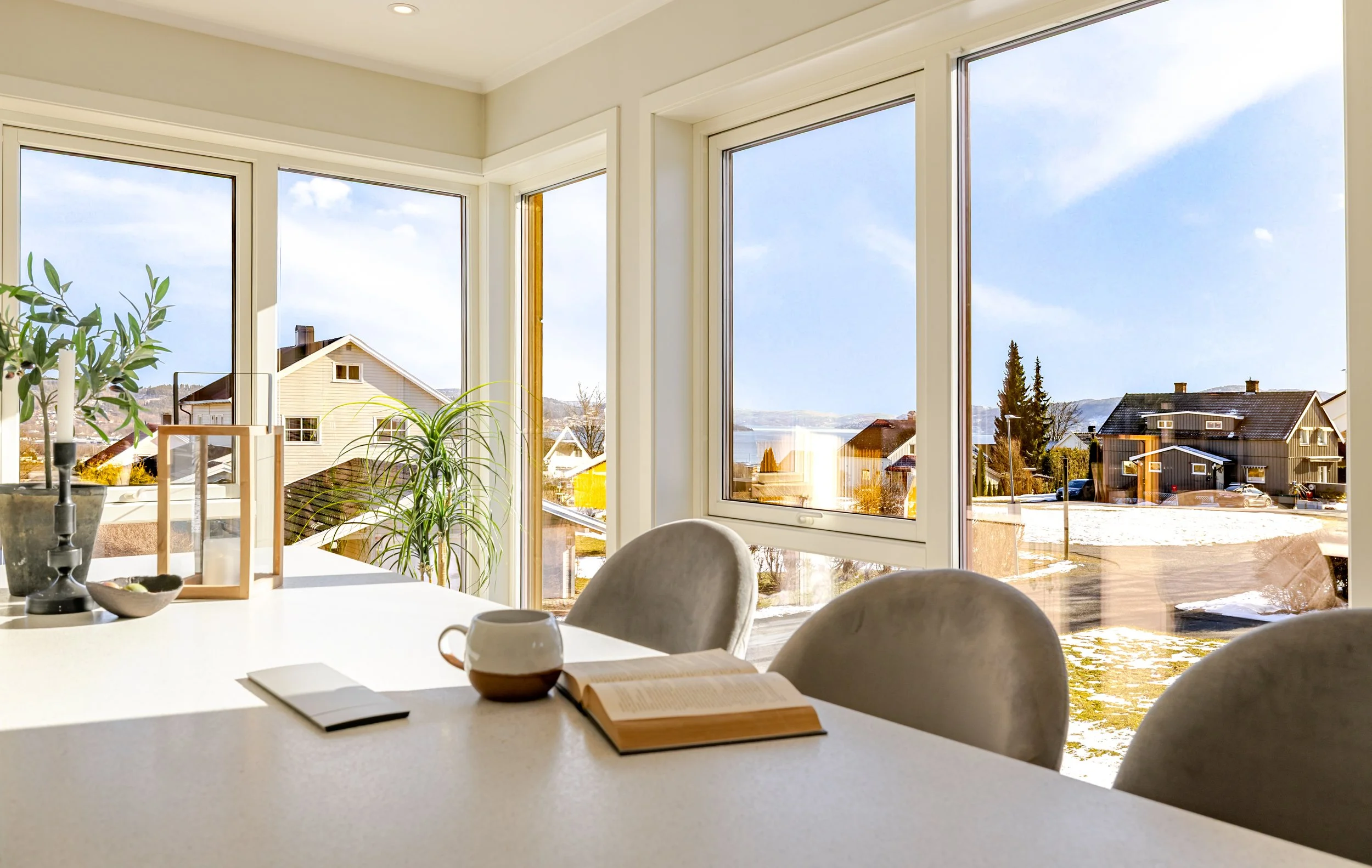 En moderne stue med lyst tregulv og lyse vegger. En lys sofa med puter står foran et stort vindu, og det er et lite bord med dekorasjoner. På venstre side er et svart metallhylle med planter og lys.
