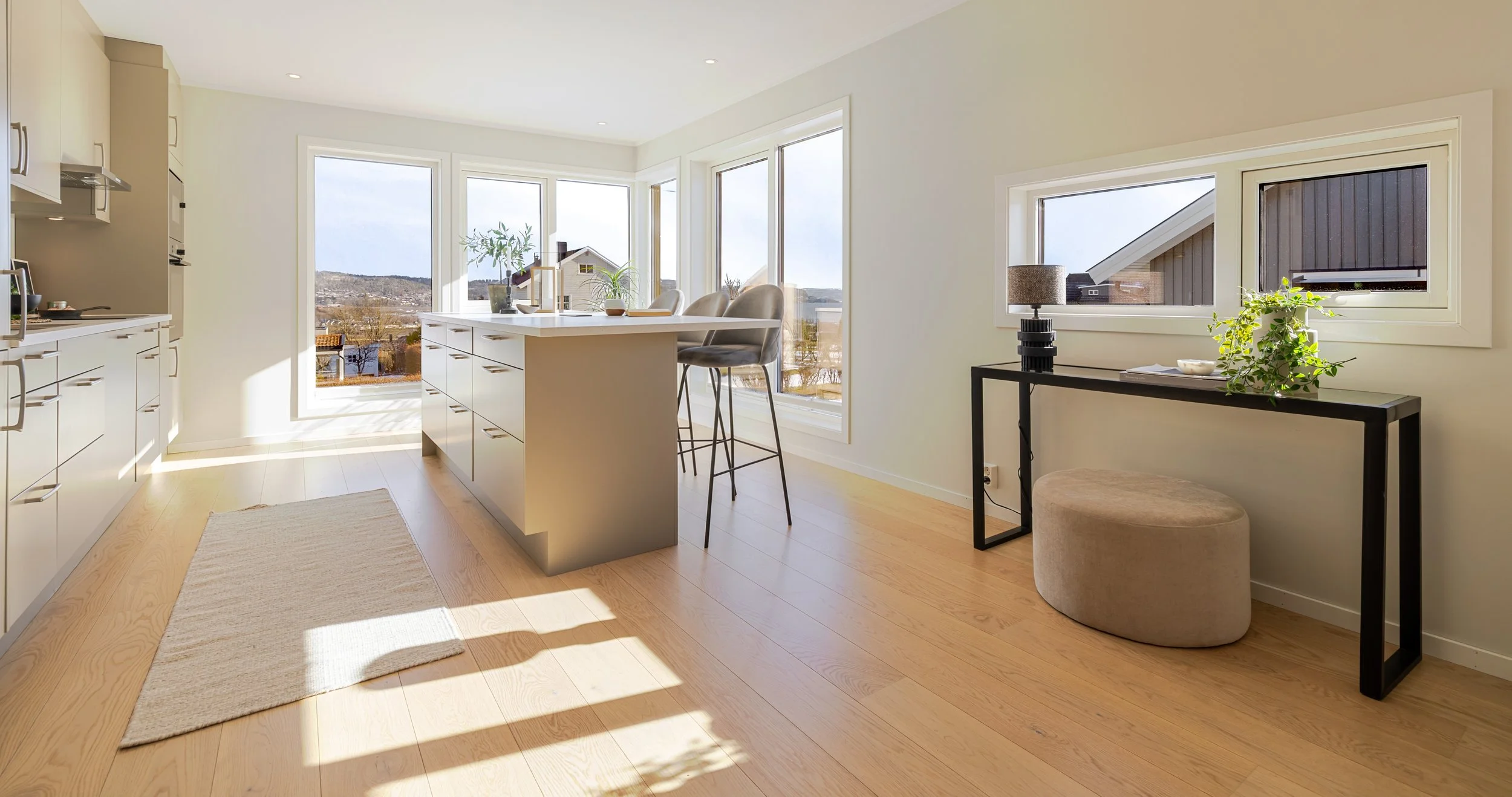 Et moderne stuebord med en blomstervase med hvite blomster, lysende stearinlys og spiseutstyr, satt opp foran store vinduer med utsikt til en terrasse og grønt landskap.