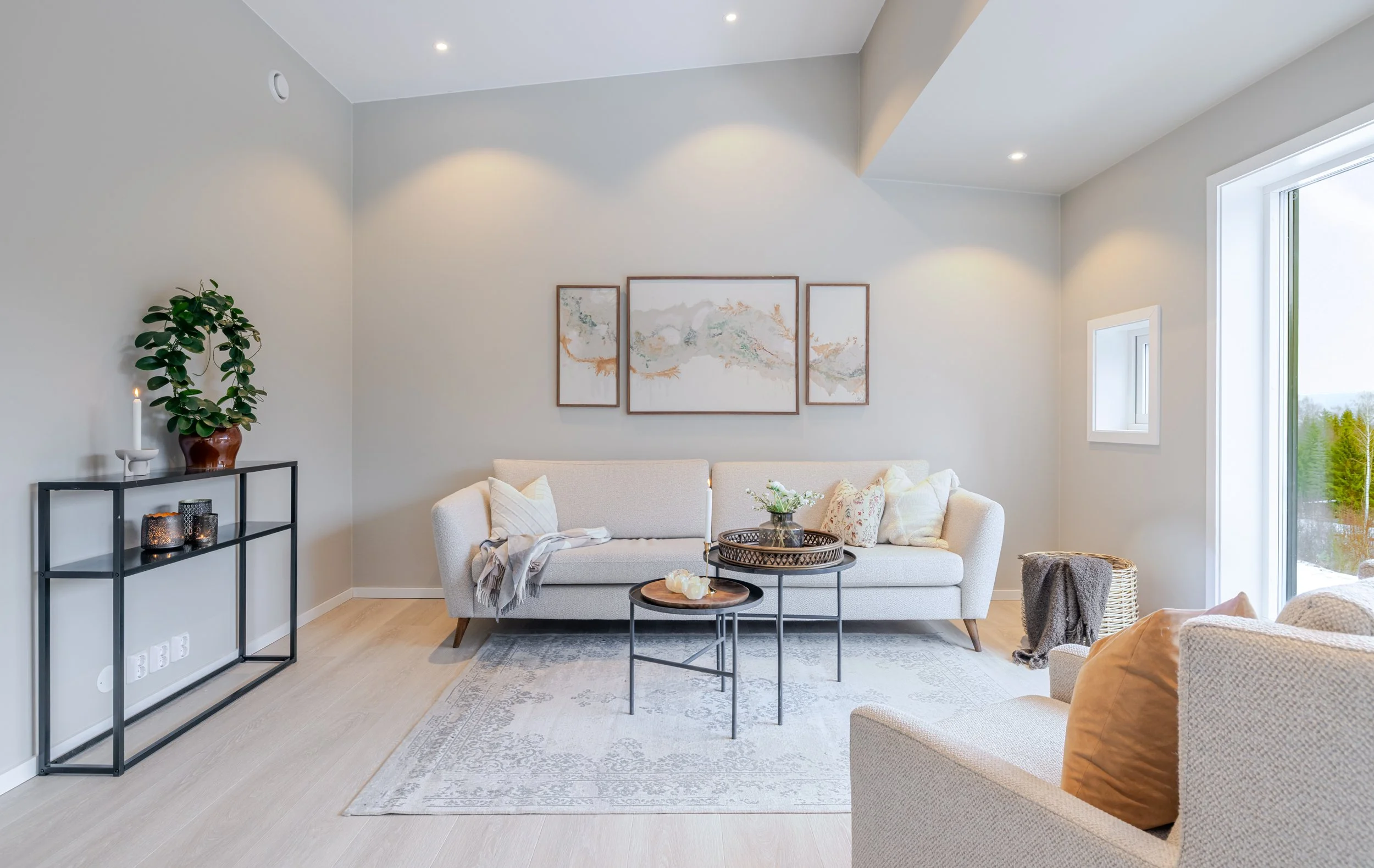 En moderne stue med lyst tregulv og lyse vegger. En lys sofa med puter står foran et stort vindu, og det er et lite bord med dekorasjoner. På venstre side er et svart metallhylle med planter og lys.
