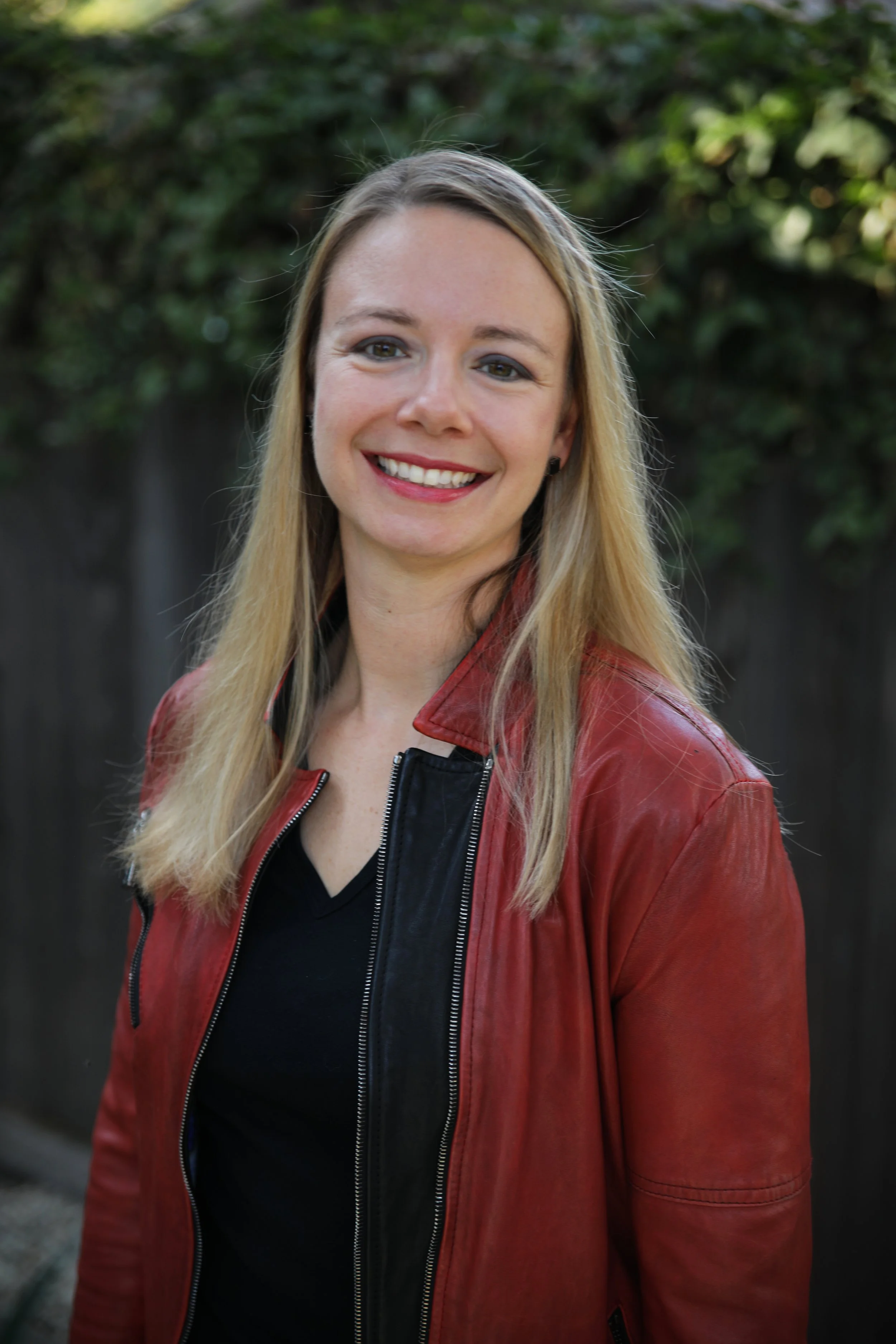 A woman with long blonde hair smiling outdoors, wearing a red leather jacket over a black top, with a blurred green foliage background.