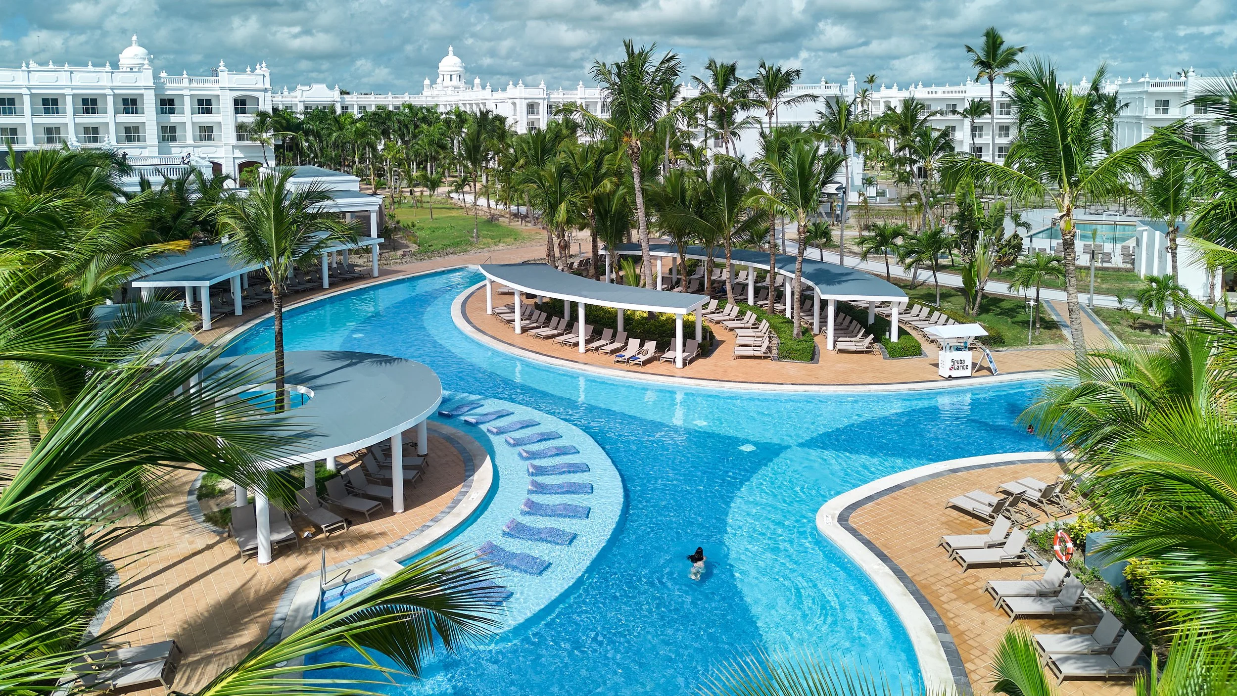 Luxury resort swimming pool with palm trees, sun loungers, shaded cabanas, and a large white building in the background.