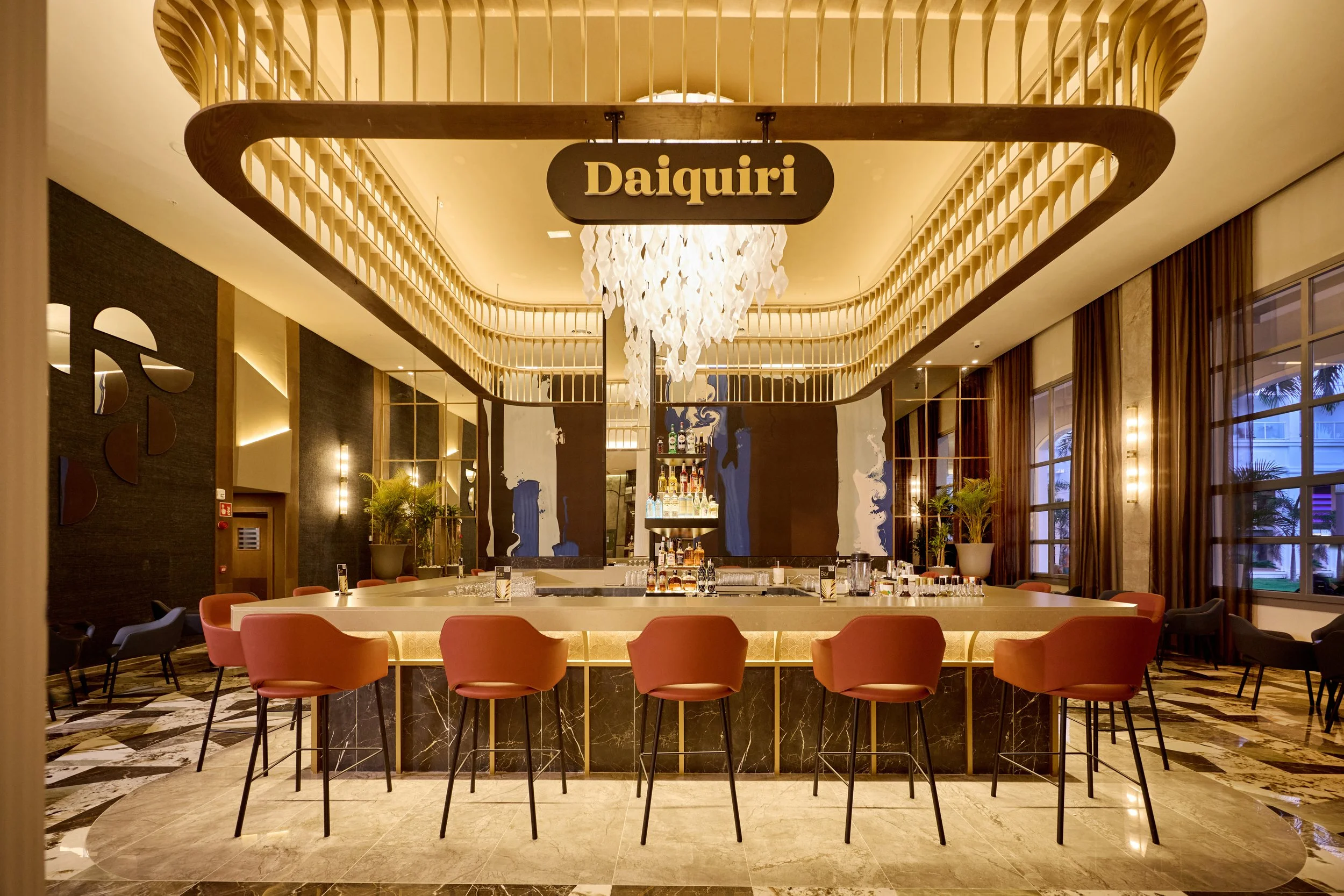 An elegant hotel bar with a circular bar counter surrounded by pink and ash-colored chairs. The bar is stocked with various bottles and glasses, with a large sign reading 'Daiquiri' hanging above. The background features large windows with brown curt
