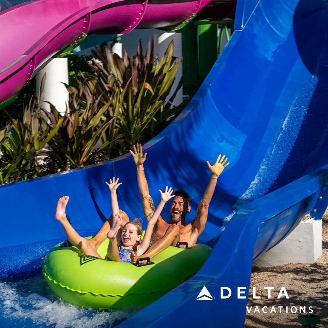 A man and a young girl riding a green inner tube down a water slide at a water park, both raising their hands and smiling. The water slide is blue with a pink and green section overhead, and there are plants beside the slide. The logo "Delta Vacation