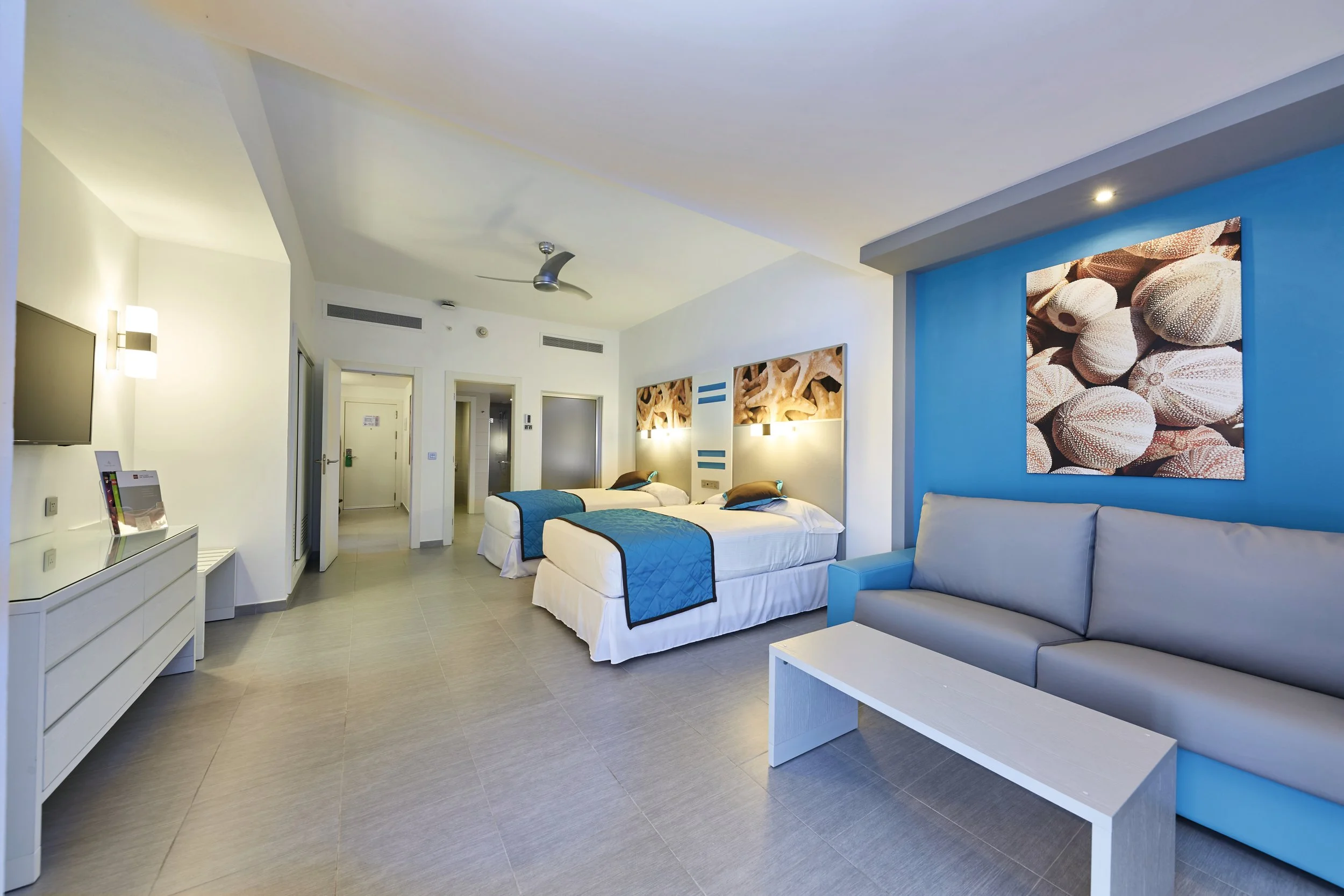 Modern hotel room with two beds, a blue accent wall with shell artwork, a gray sofa, a white coffee table, wall-mounted TV, and contemporary lighting.