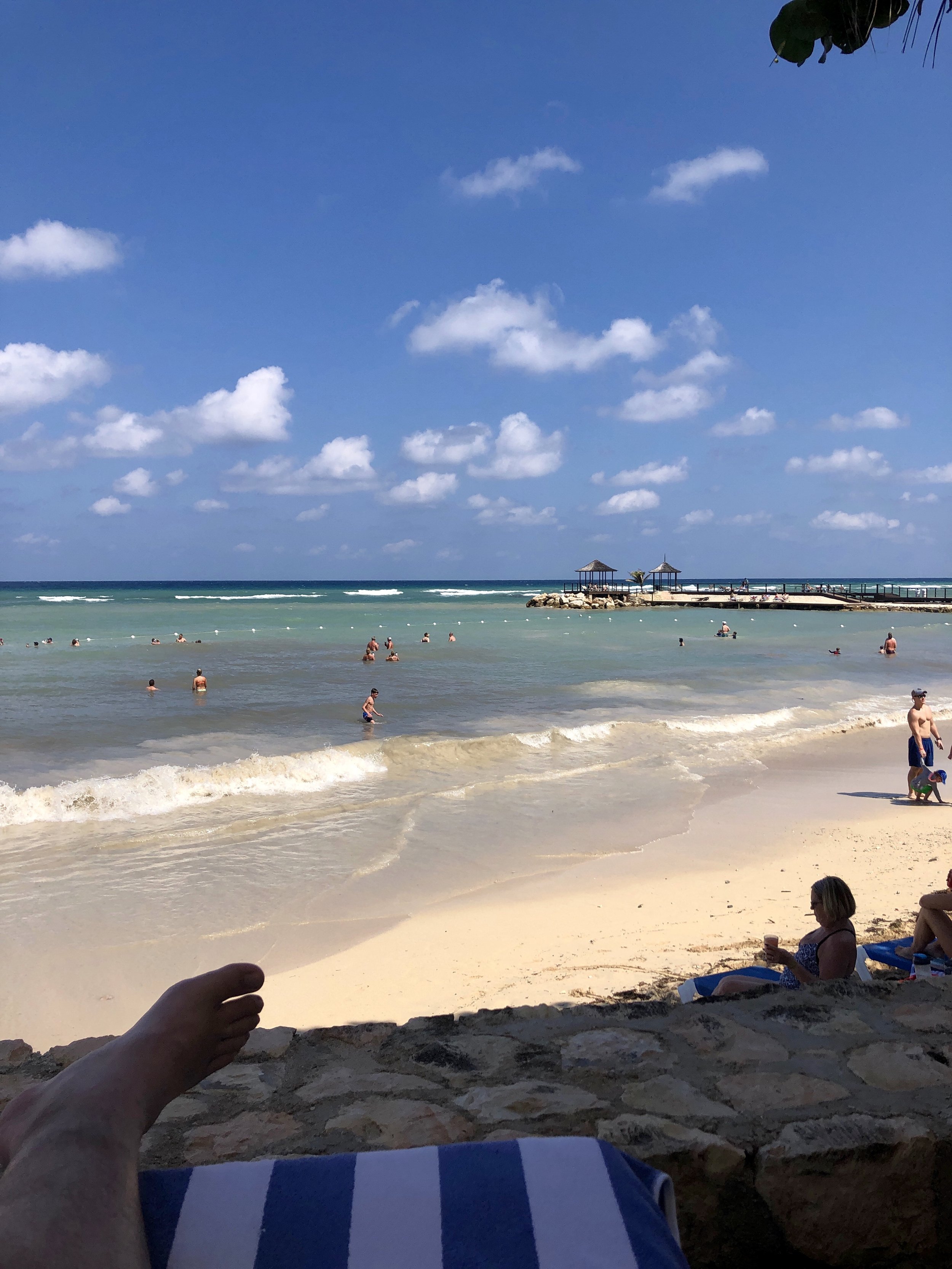 People swimming and relaxing on a beach with a rocky pier and small pavilions, under a blue sky with scattered clouds.