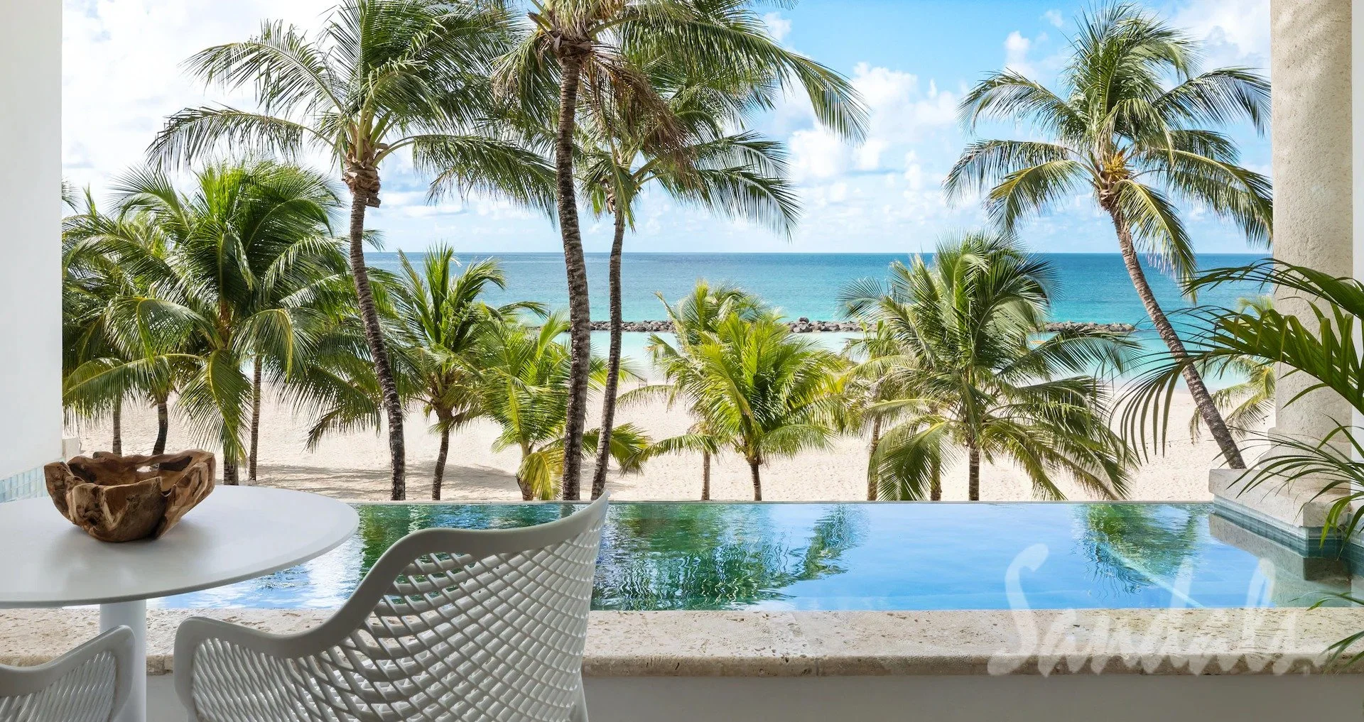 View from a balcony overlooking a tropical beach with palm trees, sandy shoreline, and turquoise ocean, partly obscured by a small pool and white table with chairs.