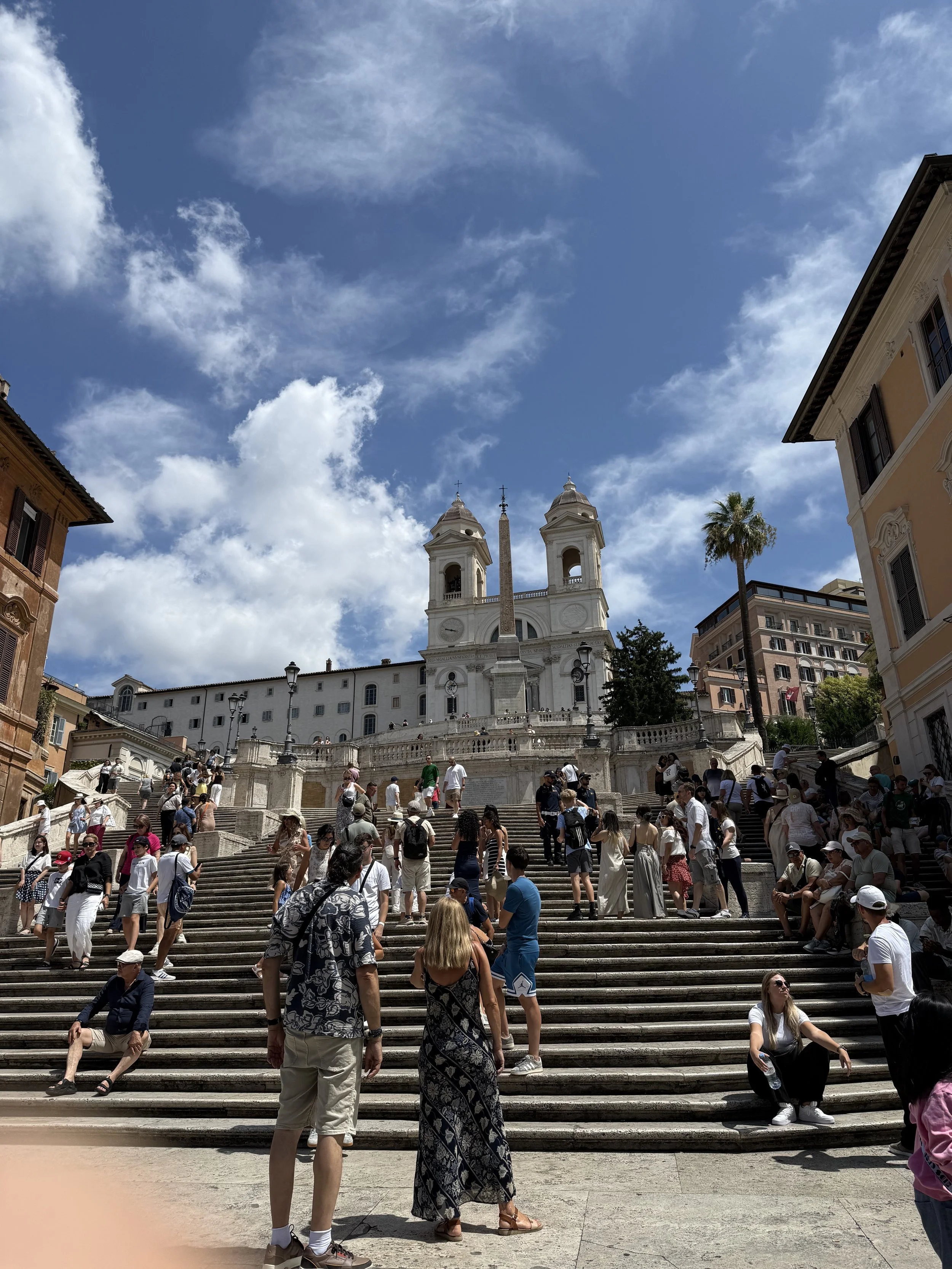 Tourists gather on the Spanish Steps leading up to the Trinità dei Monti church in Rome, Italy, under a blue sky with white clouds.