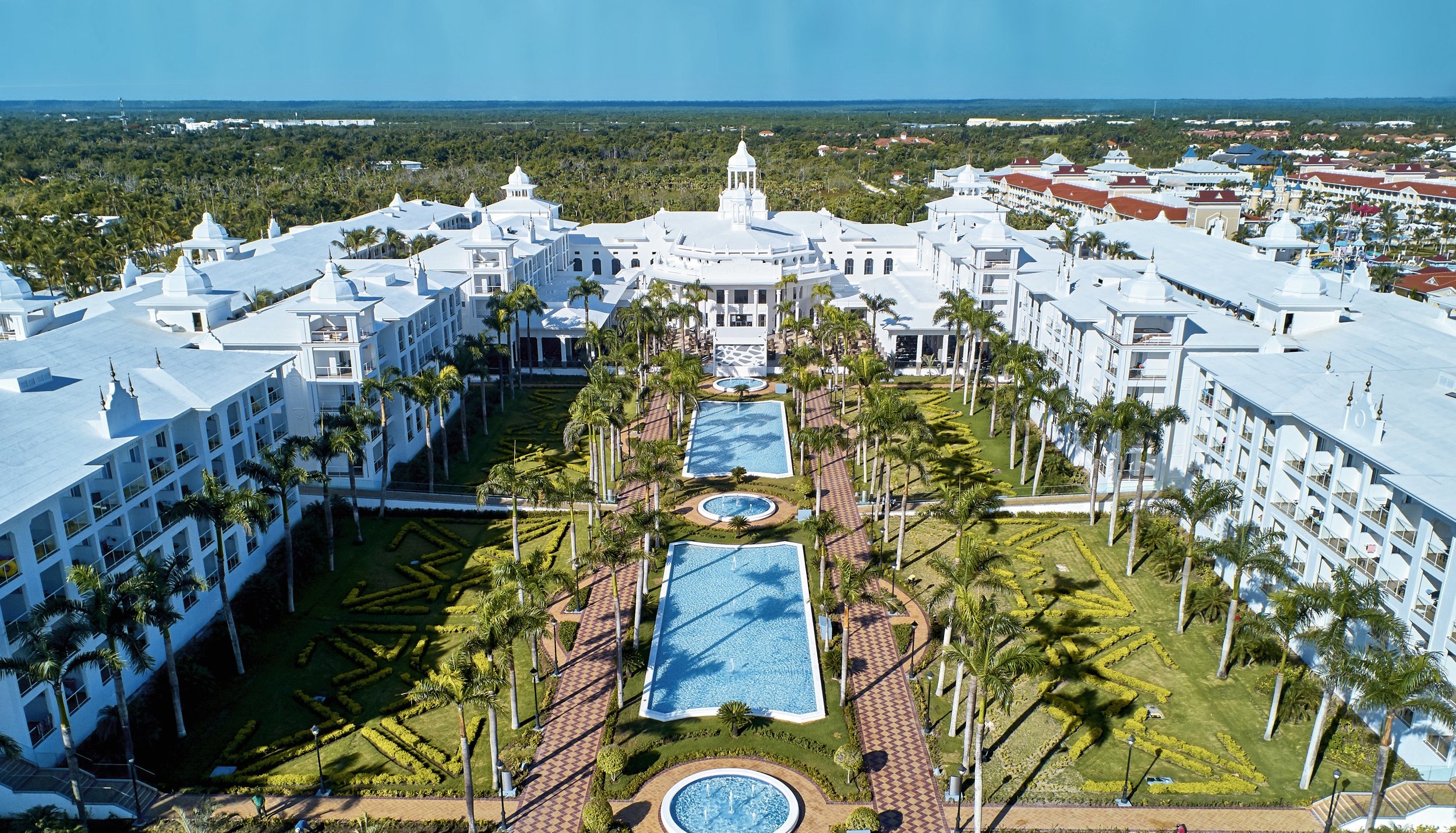 A large white hotel building with multiple spires and a central tower, surrounded by palm trees and landscaped gardens with fountains and pools, under a clear blue sky.