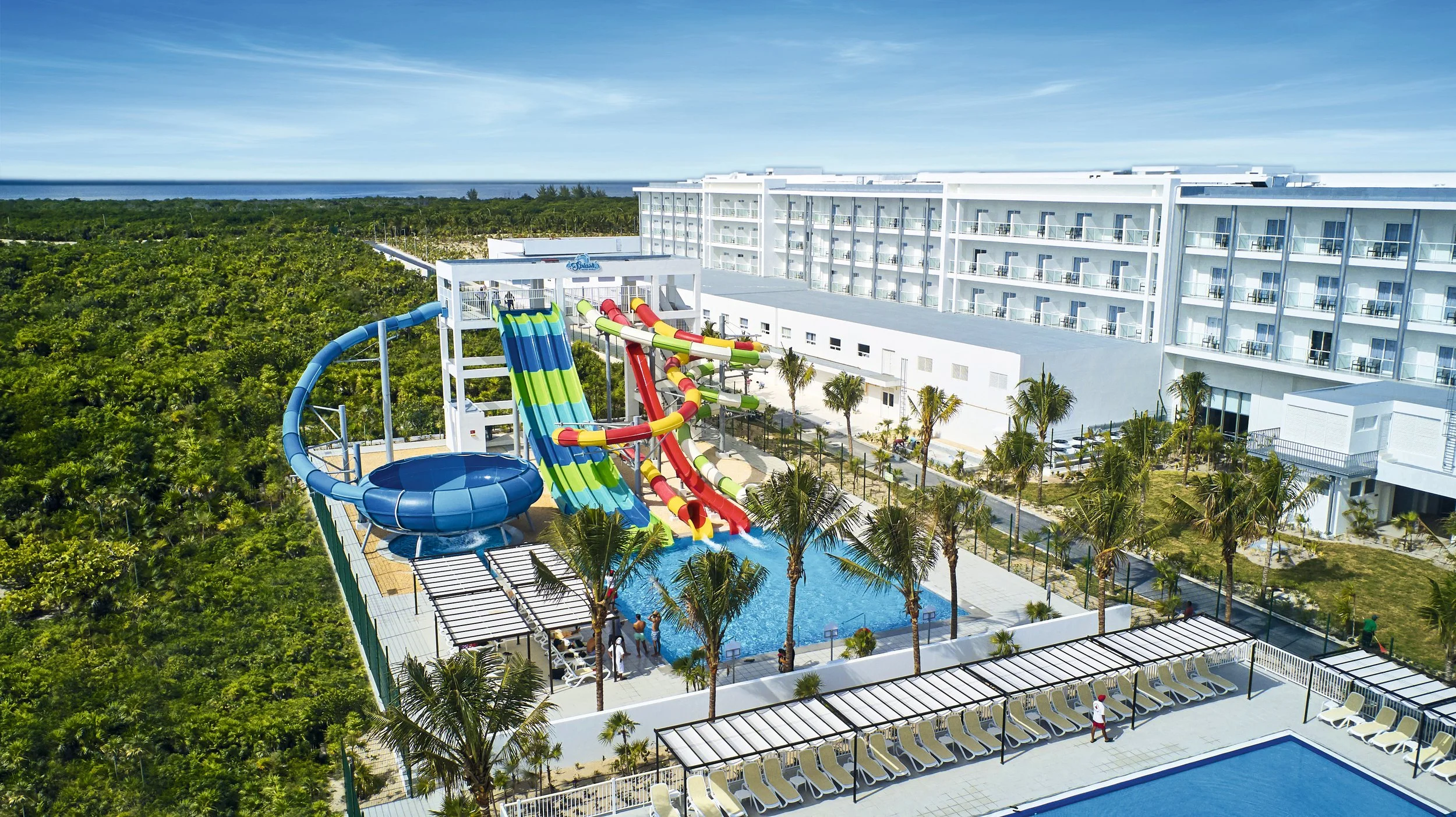 Aerial view of a water park with water slides, a pool, lounge chairs, and palm trees adjacent to a large white hotel building, with a forest and ocean in the background.