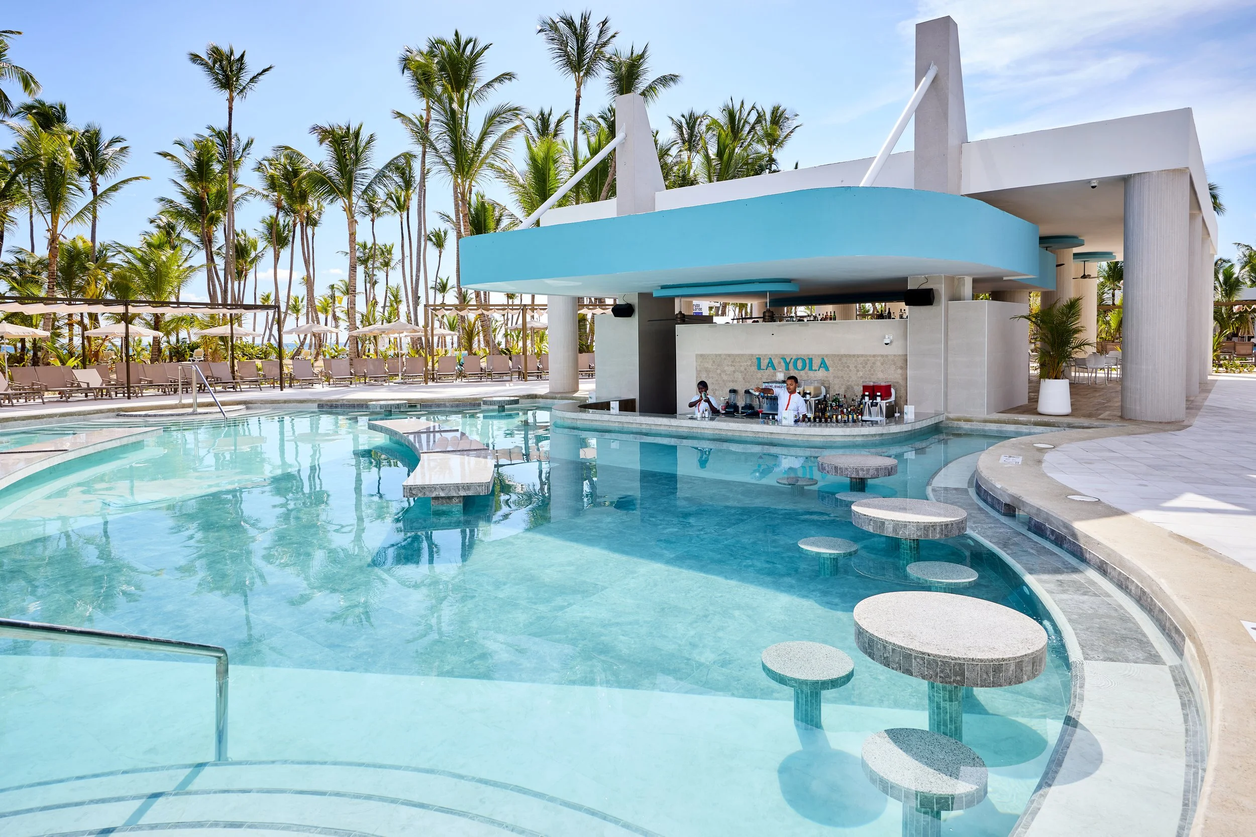 Resort pool area with a swim-up bar named La Yola, surrounded by lounge chairs, umbrellas, and palm trees.