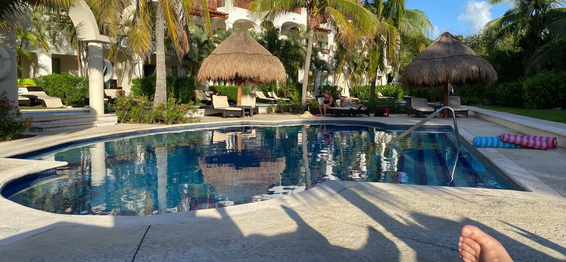 Resort pool area with lounge chairs, thatched umbrellas, palm trees, and tropical plants, with a person sitting on a lounge chair in the distance.