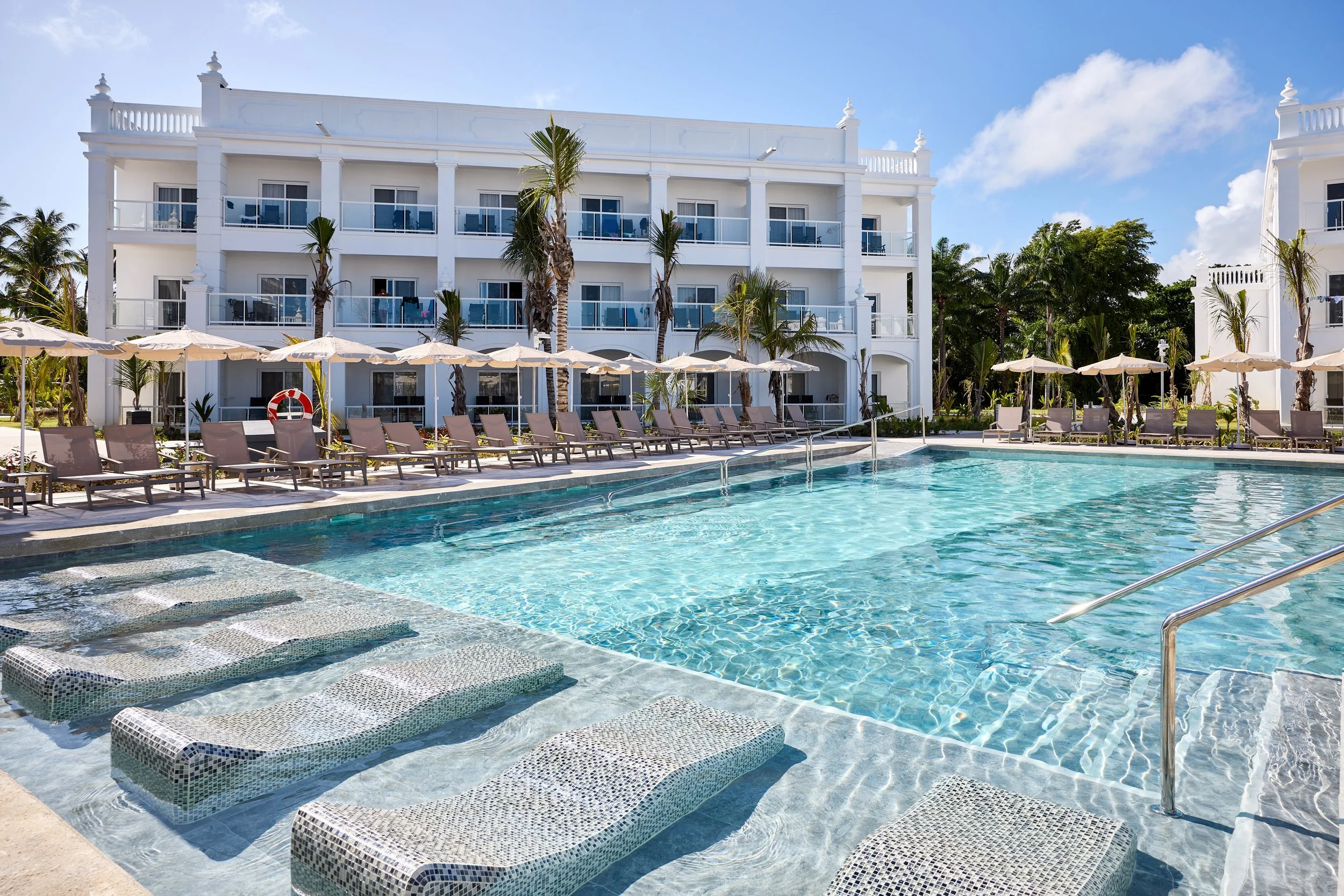 A luxurious hotel pool area with sun loungers, umbrellas, and a white multi-story hotel building in the background, surrounded by palm trees on a sunny day.