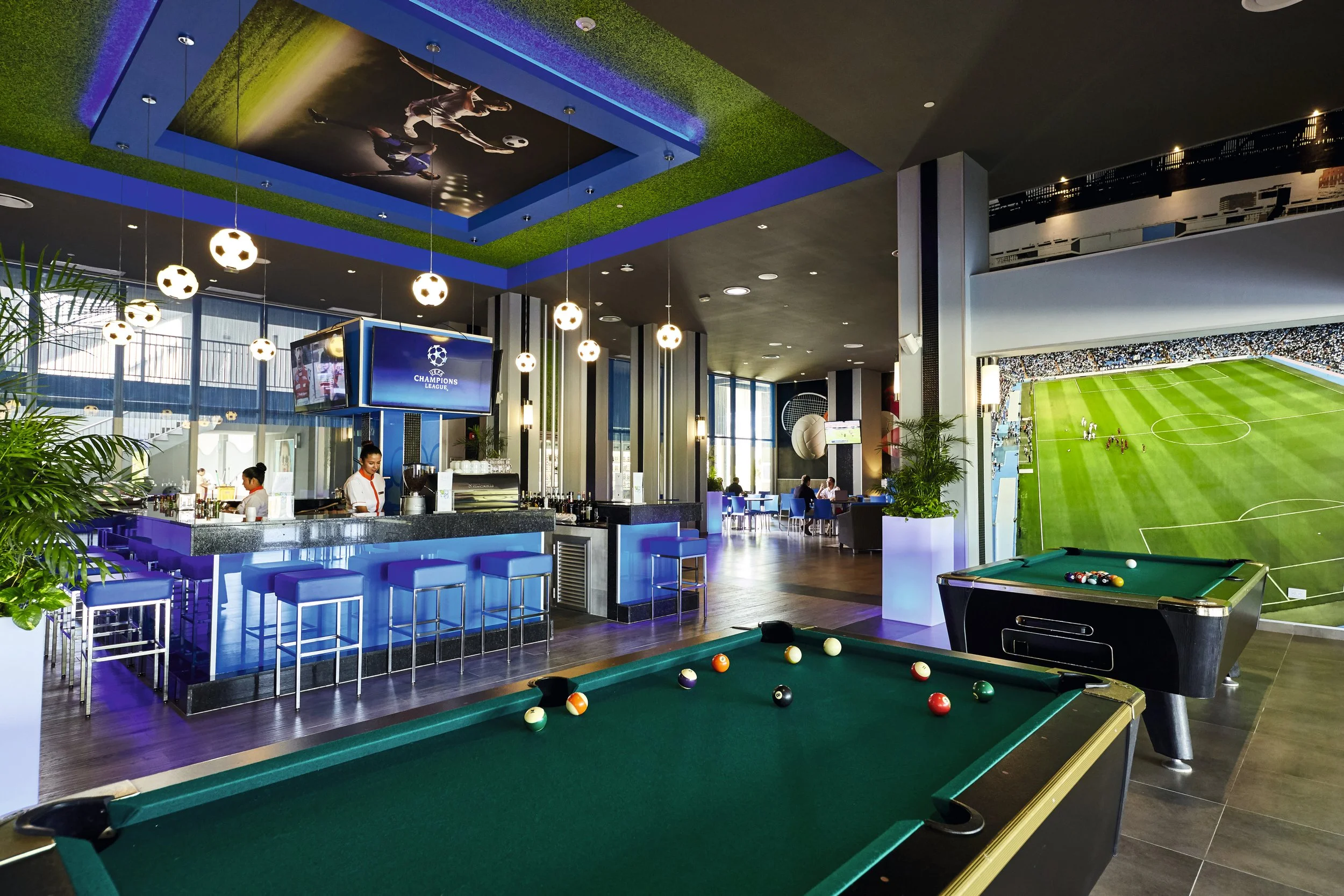 Indoor sports bar and lounge with pool tables, a bar area with high chairs, a large screen displaying a Champions League soccer match, and a panoramic viewing window of a soccer stadium.