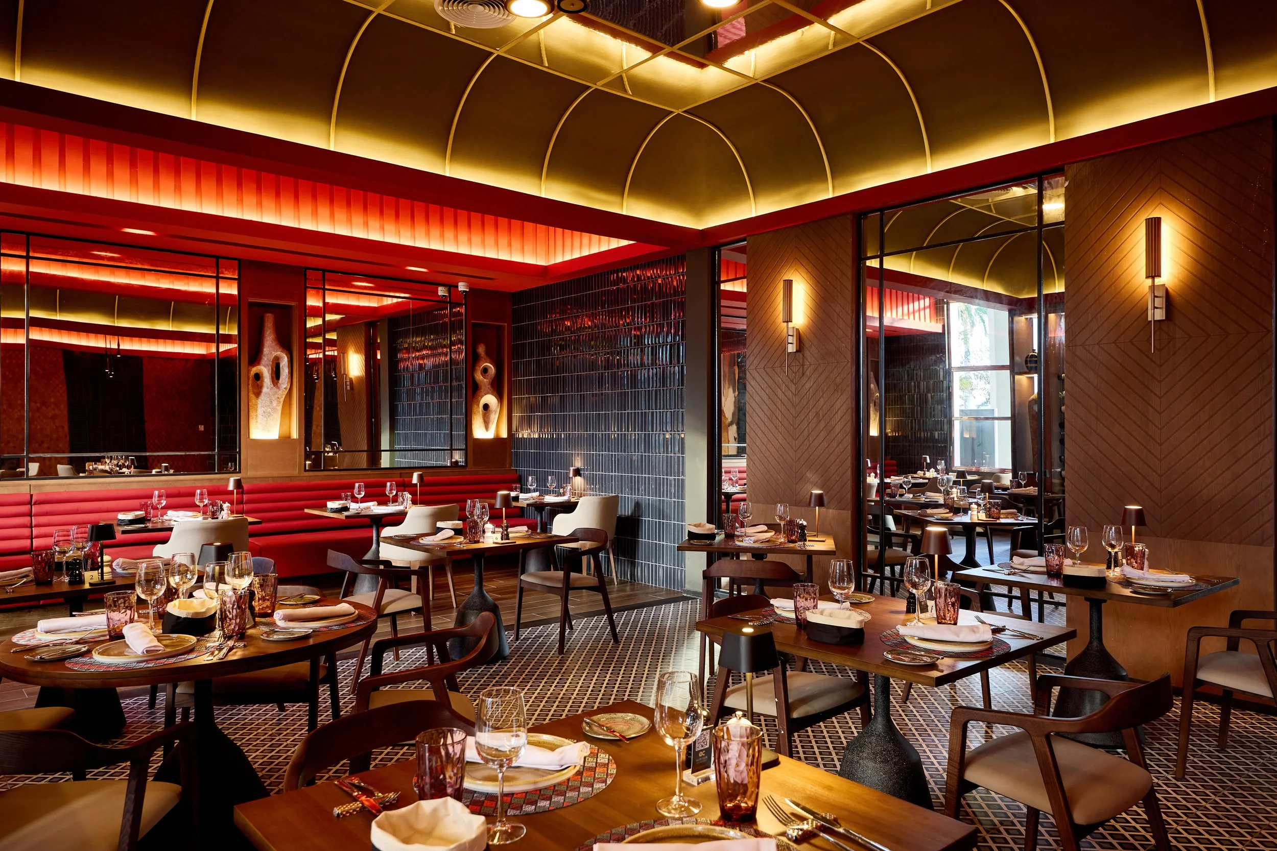 Interior of a modern upscale restaurant with tables set with wine glasses, plates, and napkins, featuring warm lighting, dark tile and wood decor, and a mirrored wall.
