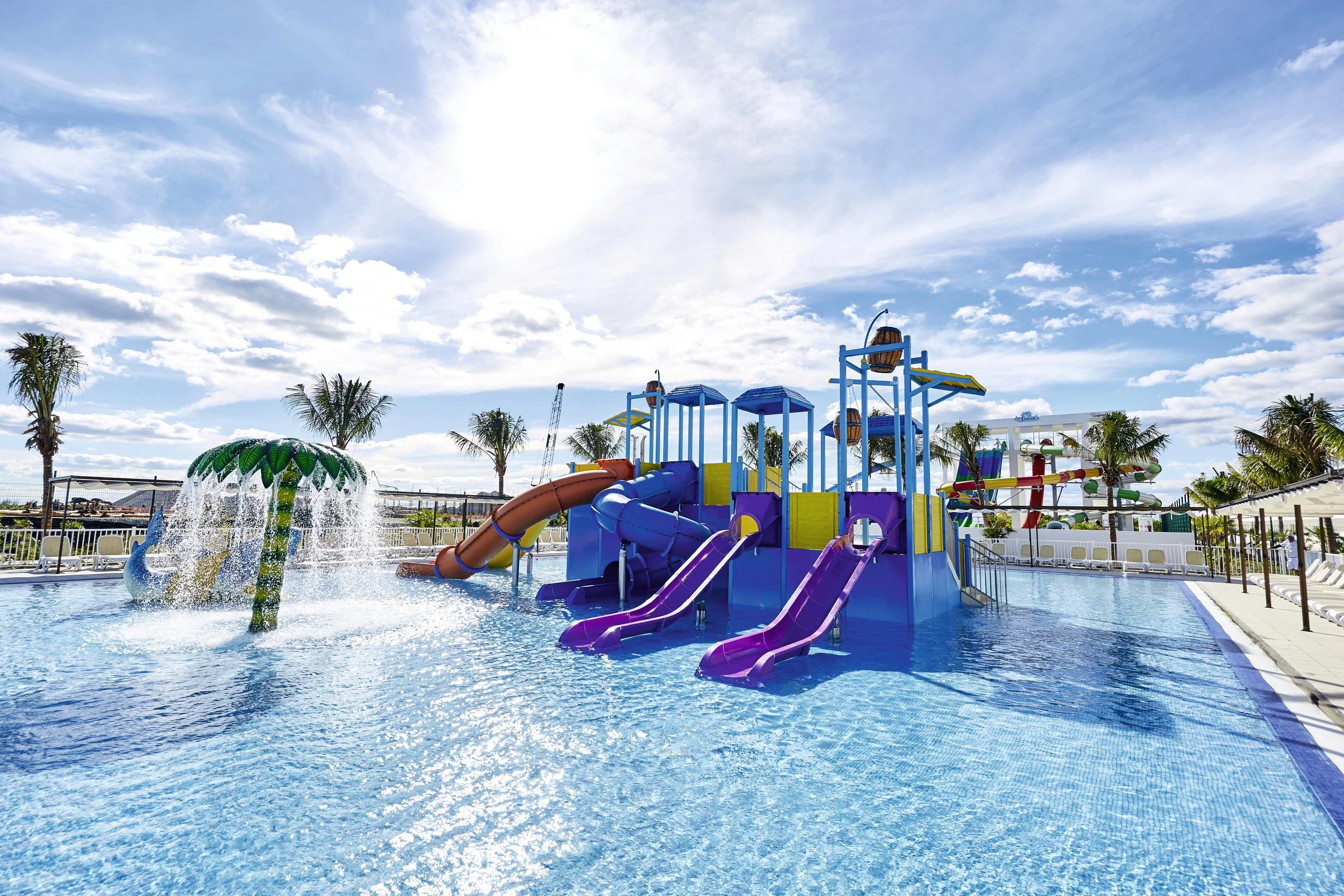 Colorful water playground with slides, a splash zone, and palm trees under a blue sky with white clouds at a pool area.