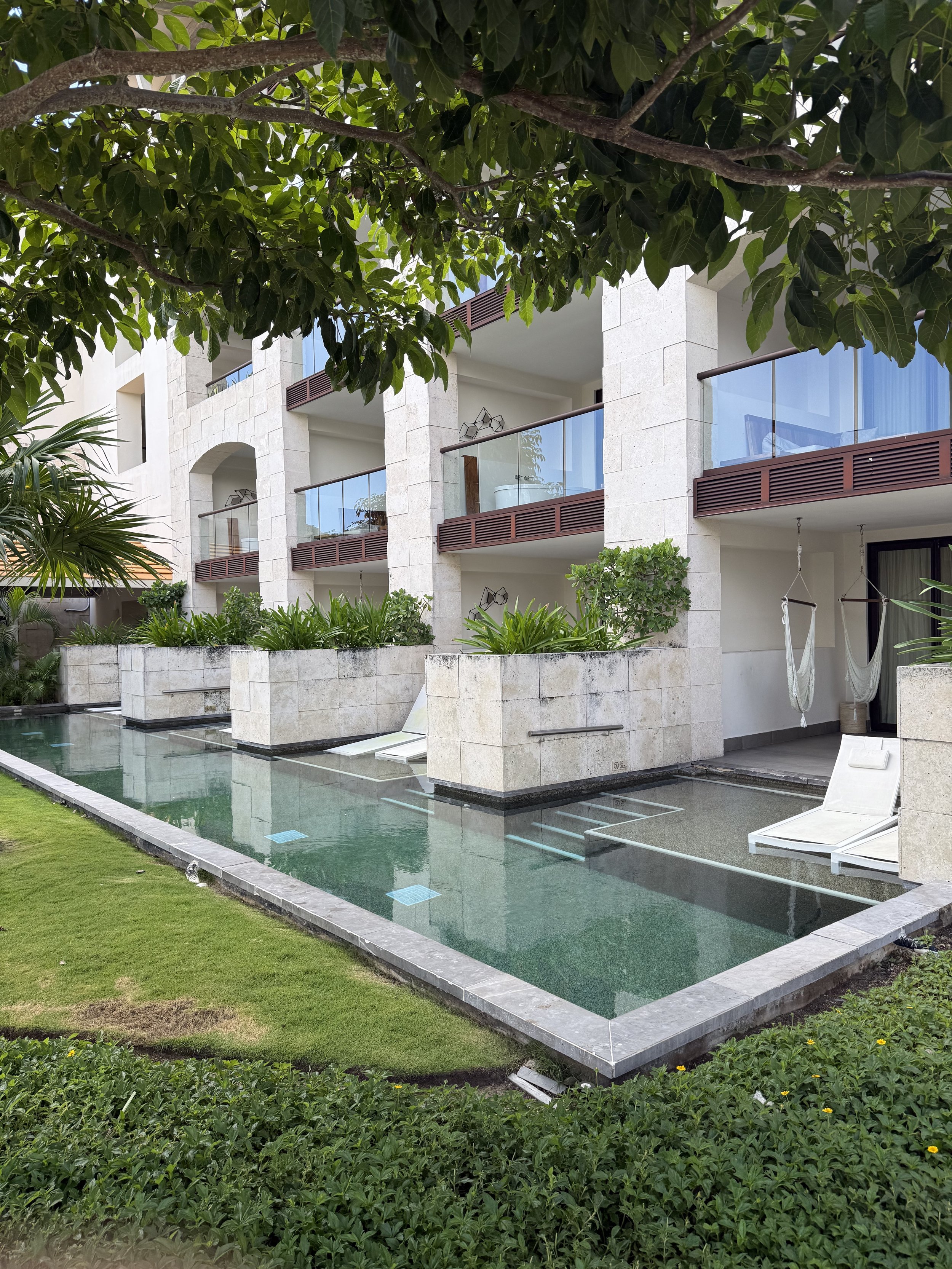 Modern apartment building with balconies, surrounded by green plants and trees, featuring a rectangular pool with submerged lounge chairs and outdoor hammocks.