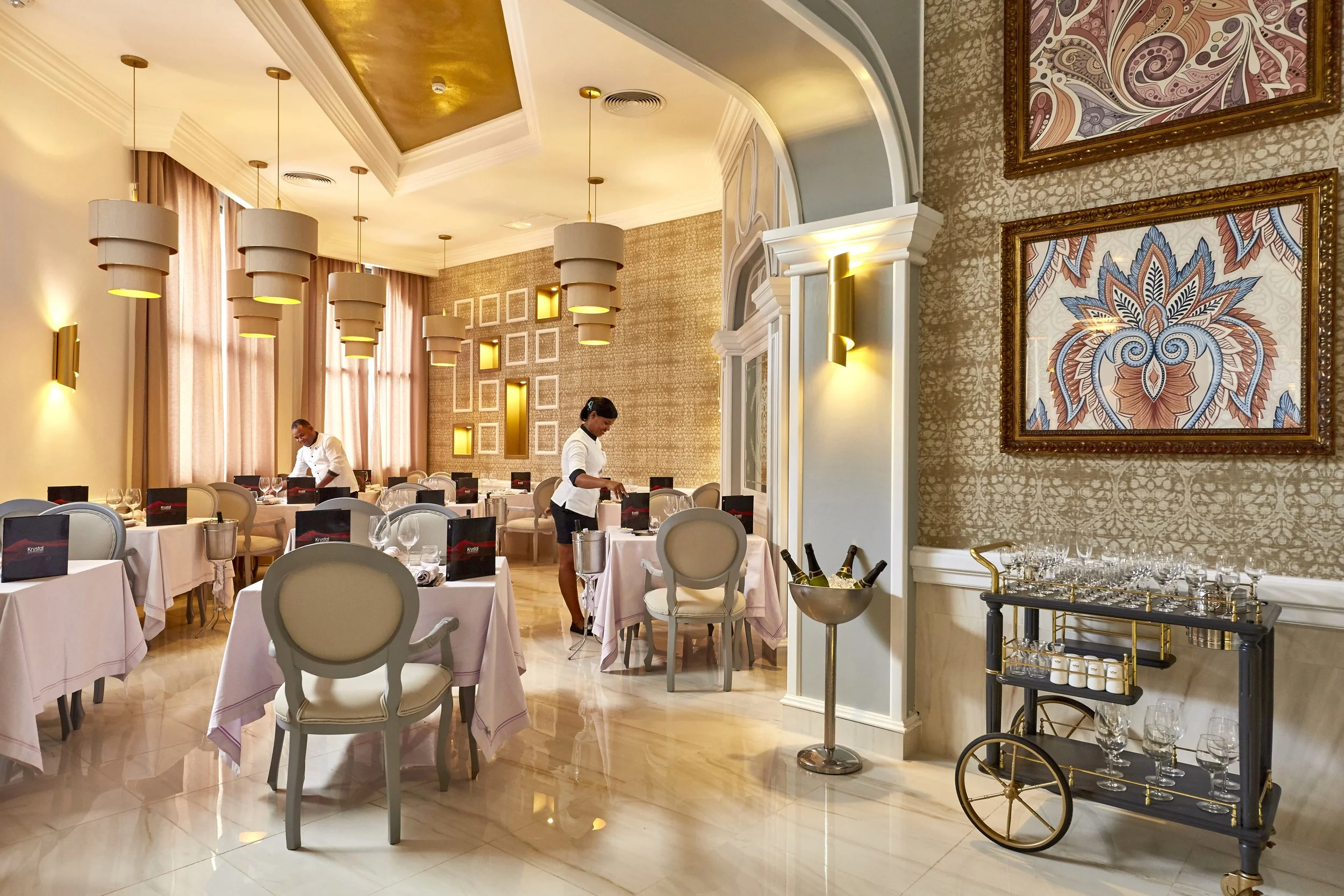 Elegant restaurant dining room with round tables covered in pink tablecloths, set with glassware and menus. Two servers preparing the tables. Modern lighting fixtures hanging from the ceiling and decorative wall art. Champagne bottles on a cart near 