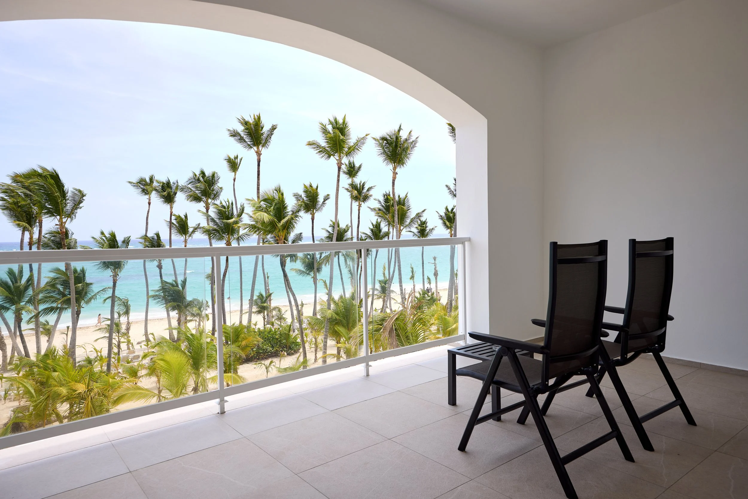 Balcony overlooking a tropical beach with palm trees and ocean view, two black chairs facing outward.
