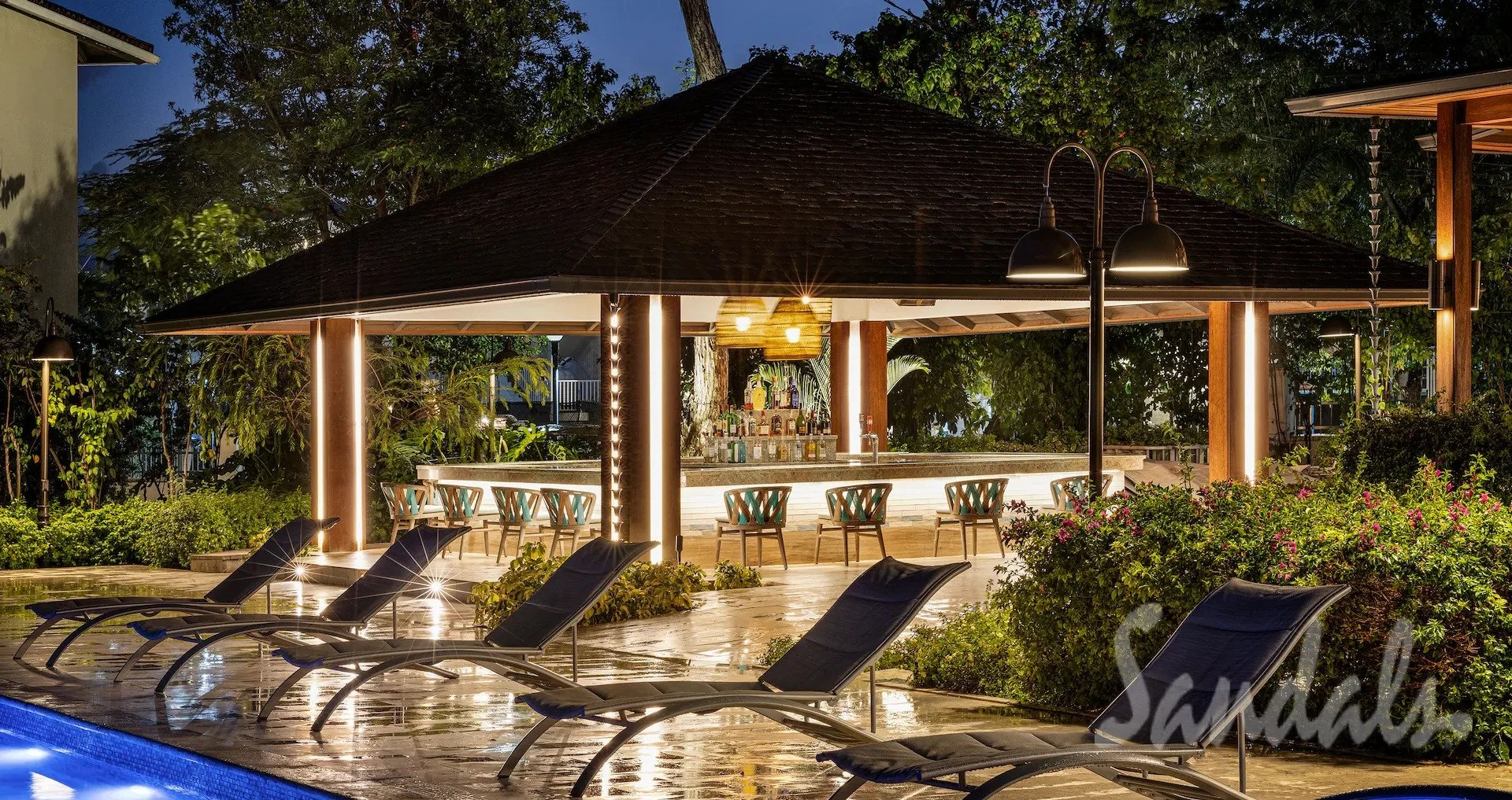 Nighttime view of an outdoor bar with bar stools, surrounded by poolside lounge chairs, greenery, and trees, illuminated with warm lighting.
