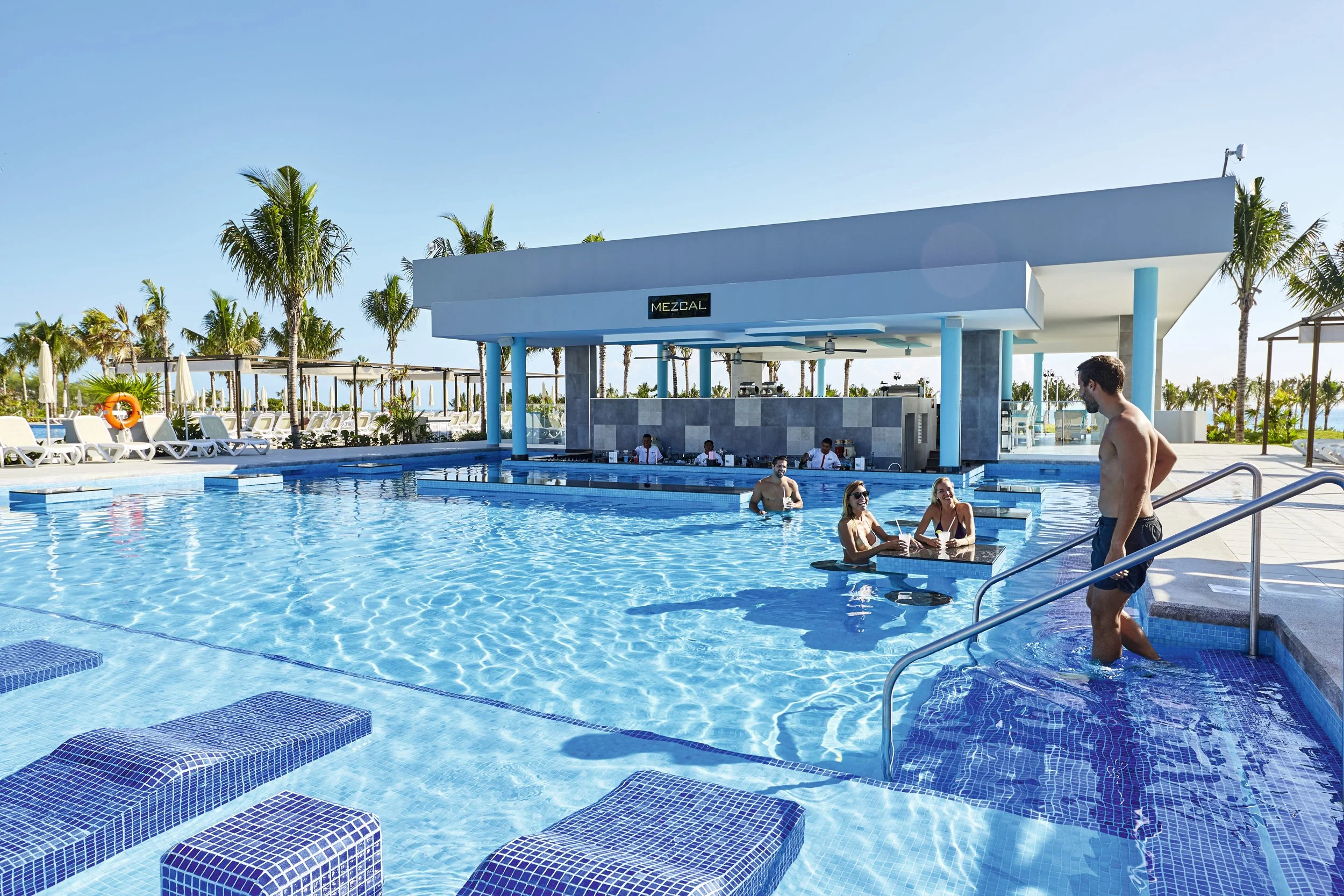 People enjoying a poolside bar at a resort with palm trees and lounge chairs, under a clear blue sky.