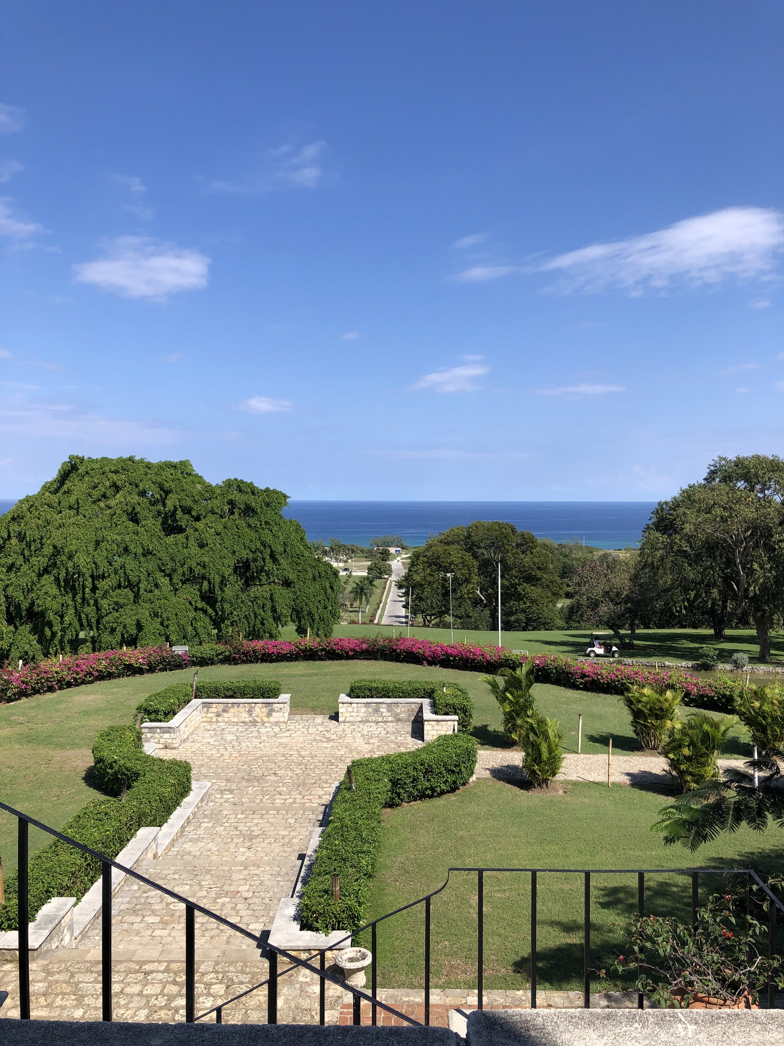 Scenic view of a garden with a stone pathway, lush green trees, pink flowering bushes, and a distant ocean under a clear blue sky.
