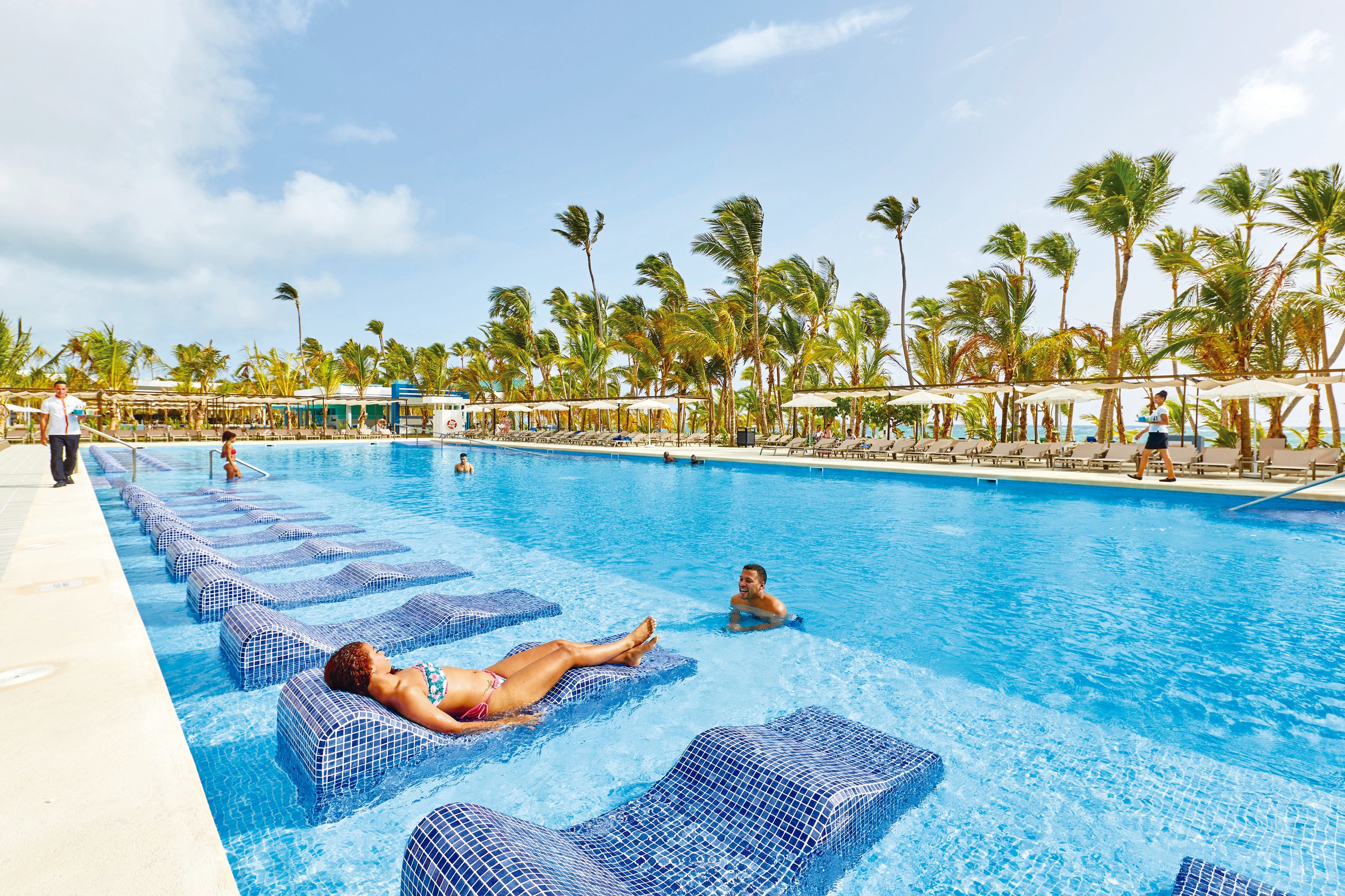 People enjoying a sunny day at a large outdoor swimming pool with palm trees and lounge chairs along the side.