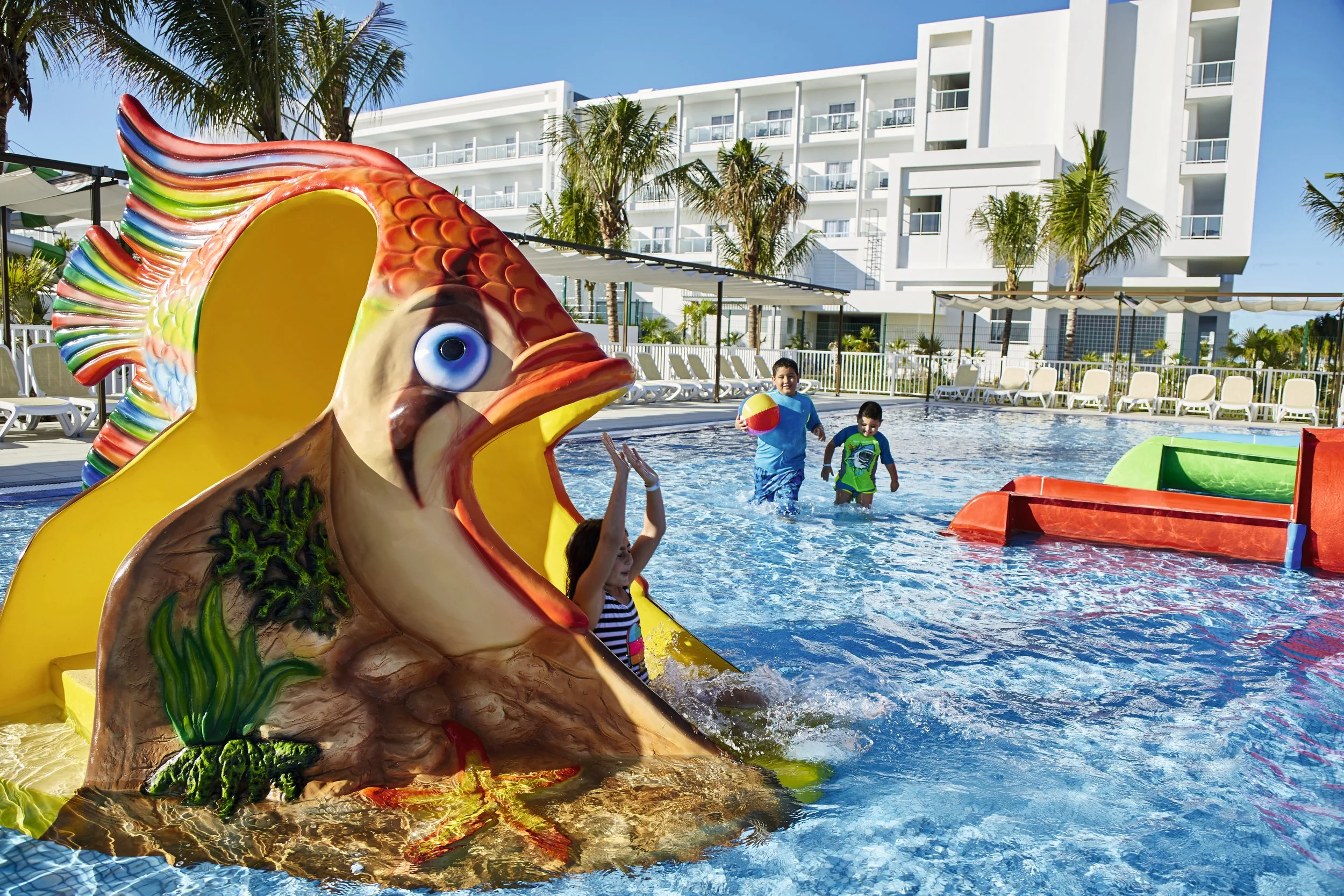 Children playing in a swimming pool with a large colorful fish slide, surrounded by lounge chairs and palm trees, with a modern hotel in the background.