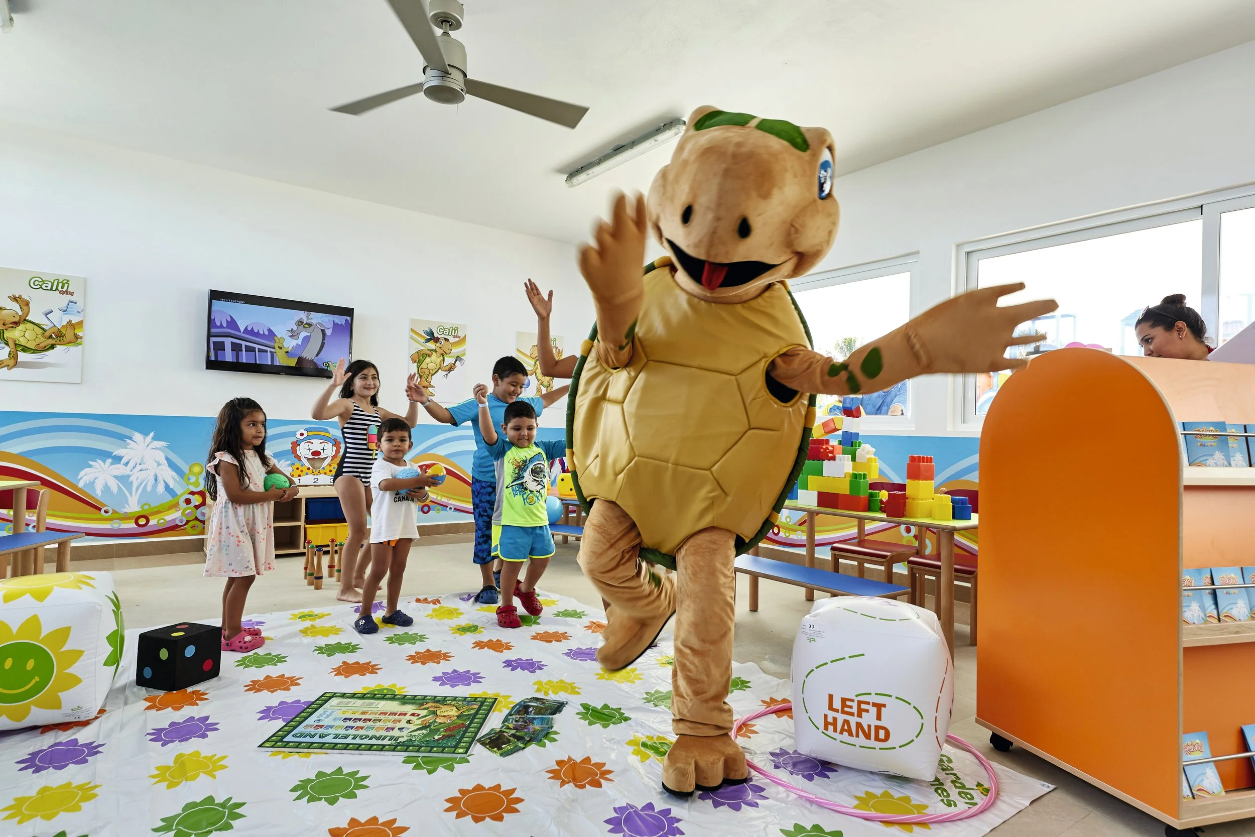Children at a birthday party or celebration with a person dressed in a turtle mascot costume. The mascot is in the middle of a game or activity on a colorful mat with flowers, while some children are watching and others are participating. The room ha