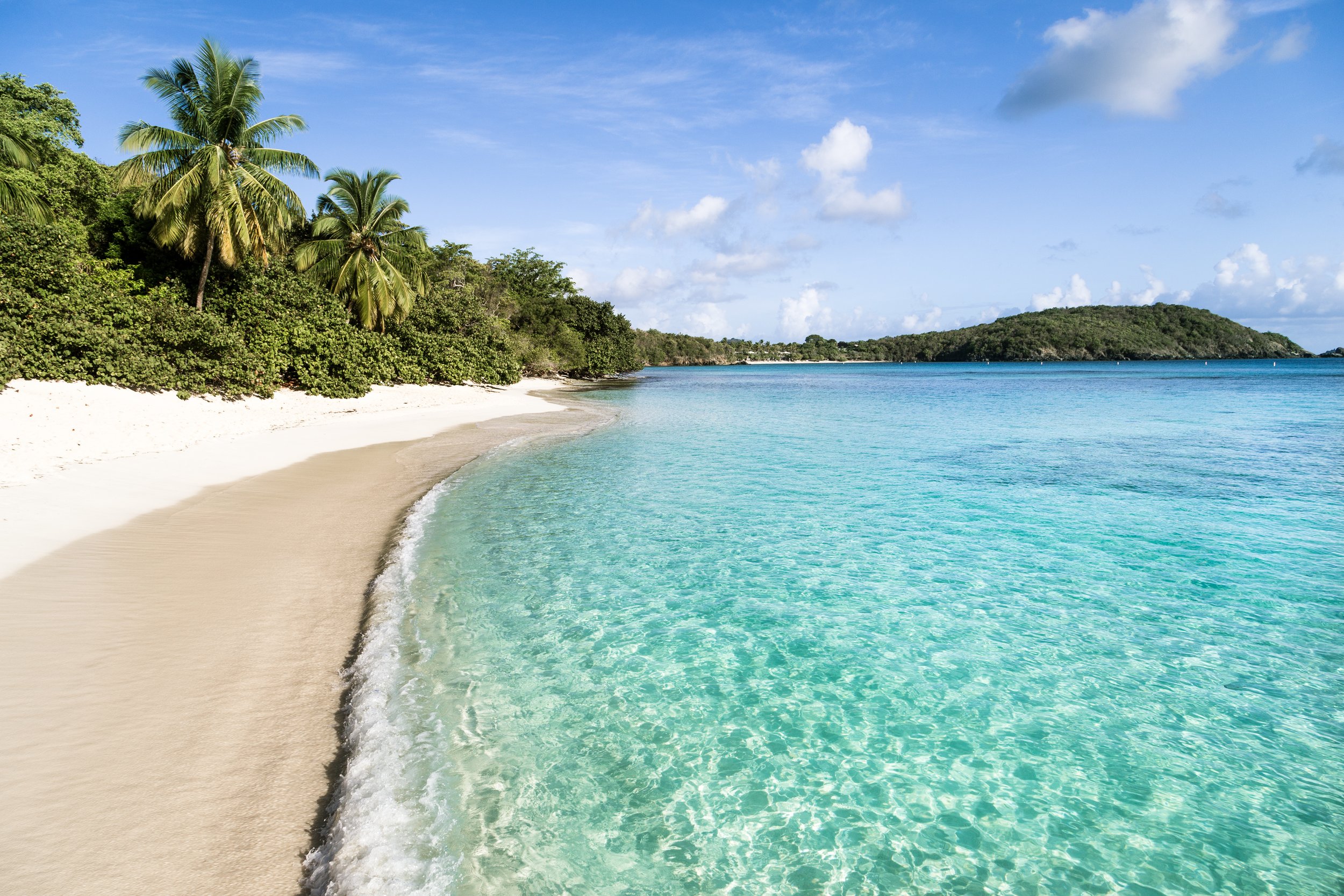 Tropical beach with white sand, turquoise water, lush green trees including palm trees, and a small island in the distance under a partly cloudy sky.