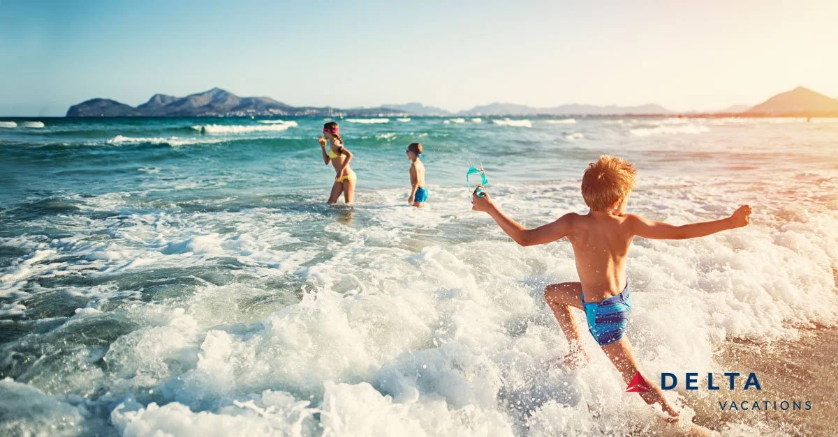 Children playing in the ocean at the beach with mountains in the background during sunset, with a "Delta Vacations" logo in the bottom right corner.