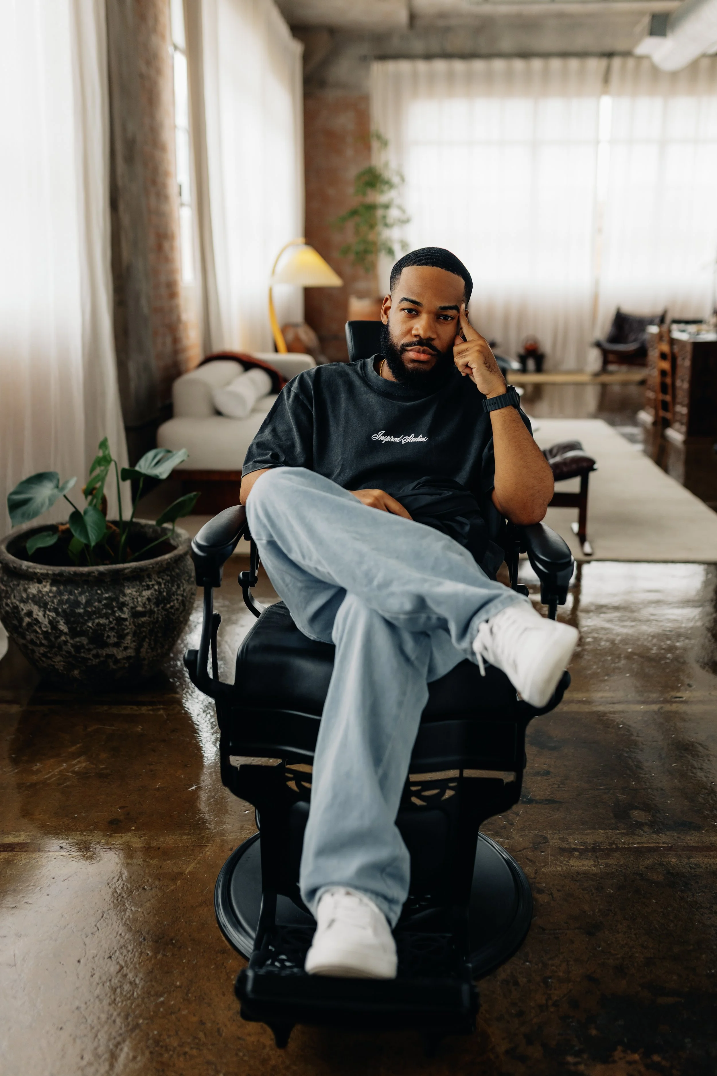 A man sitting in a black leather chair in a cozy living room with exposed brick walls, large windows with sheer curtains, a beige sofa, a yellow lamp, and potted plants.