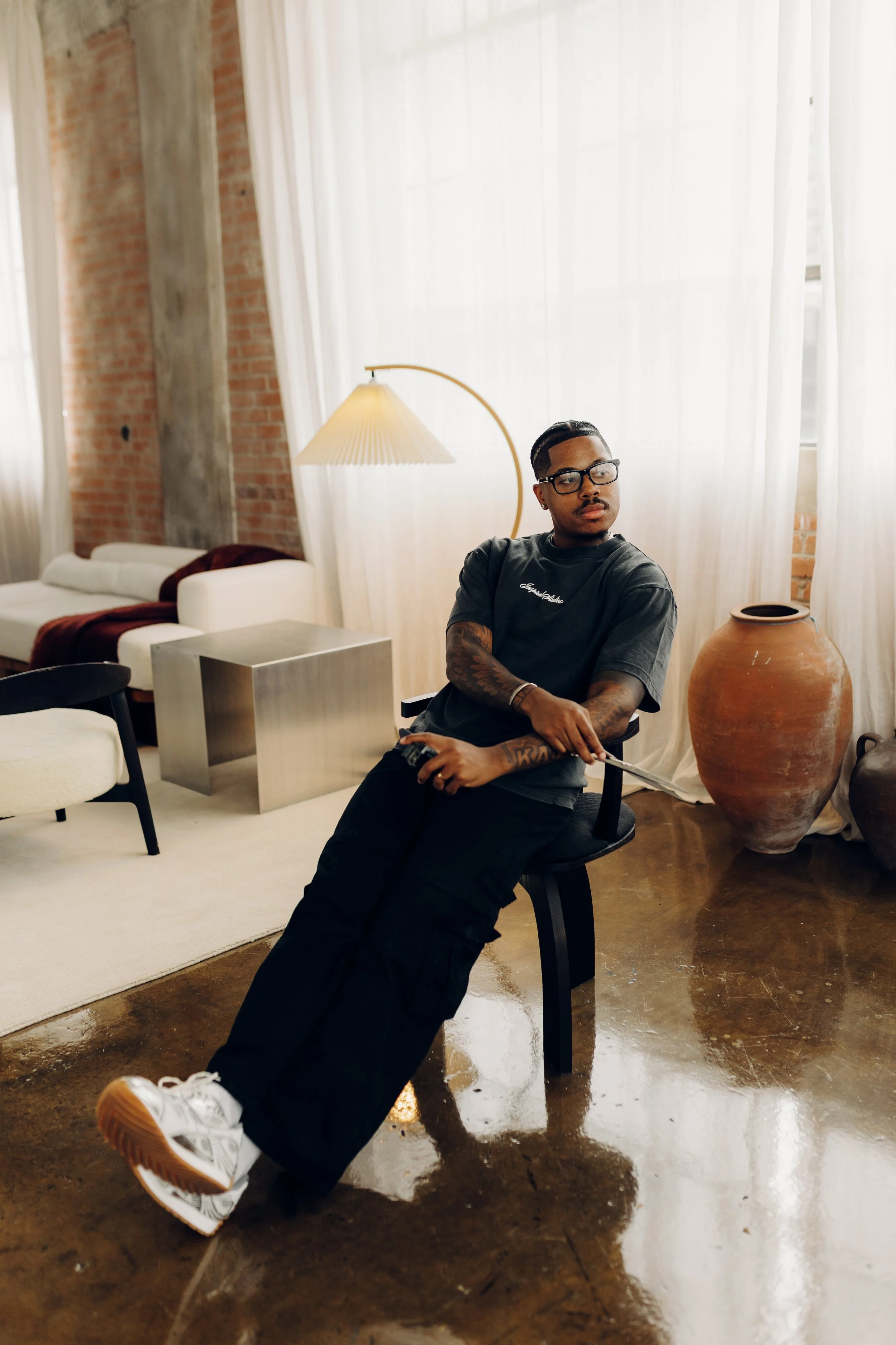 A young man with tattoos, wearing glasses, a black t-shirt, and black cargo pants, seated on a black chair in a modern living room with large windows, white curtains, a large floor lamp, and decorative ceramic pots.