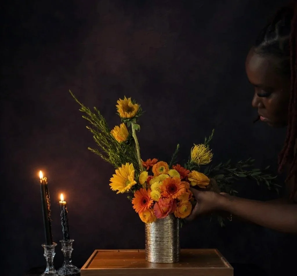 A woman arranging a vibrant bouquet of yellow, orange, and red flowers in a metallic vase on a wooden table, with two lit candles in crystal holders on the left side, against a dark background.