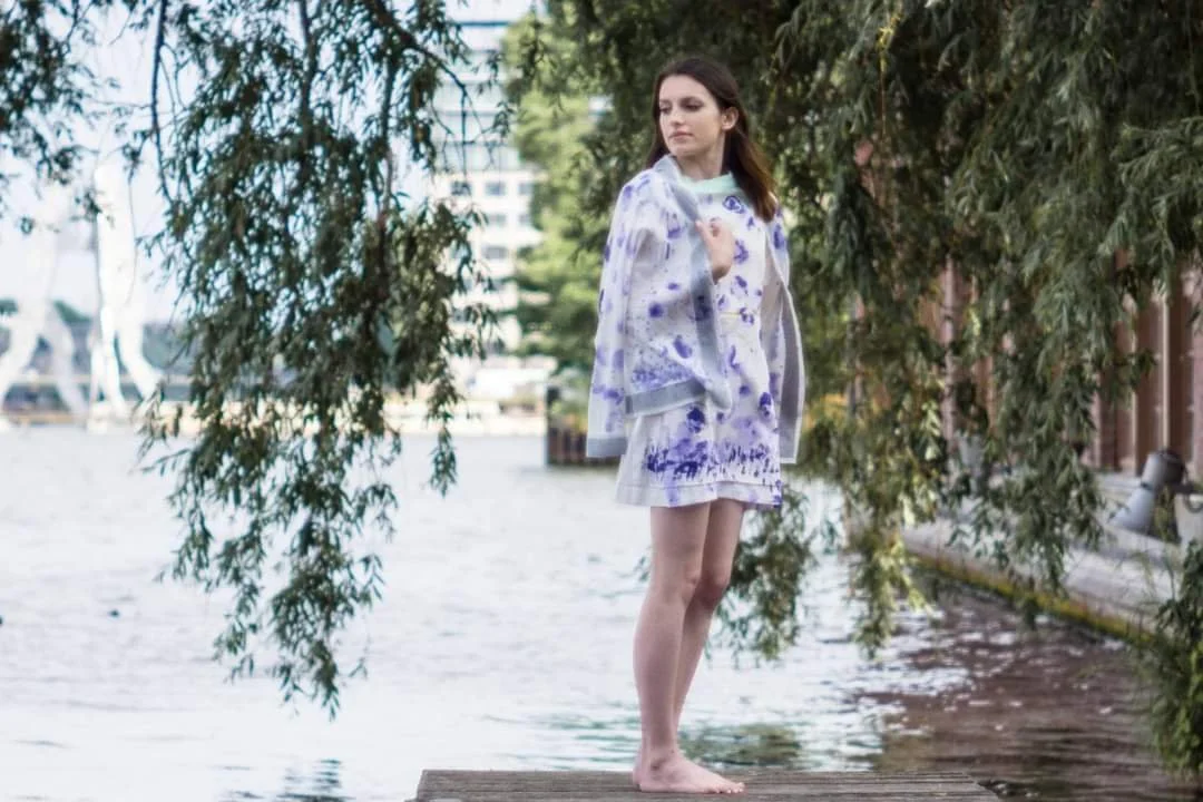 A young woman standing barefoot on a wooden dock near water, holding a patterned jacket over her shoulder, with trees and buildings in the background.