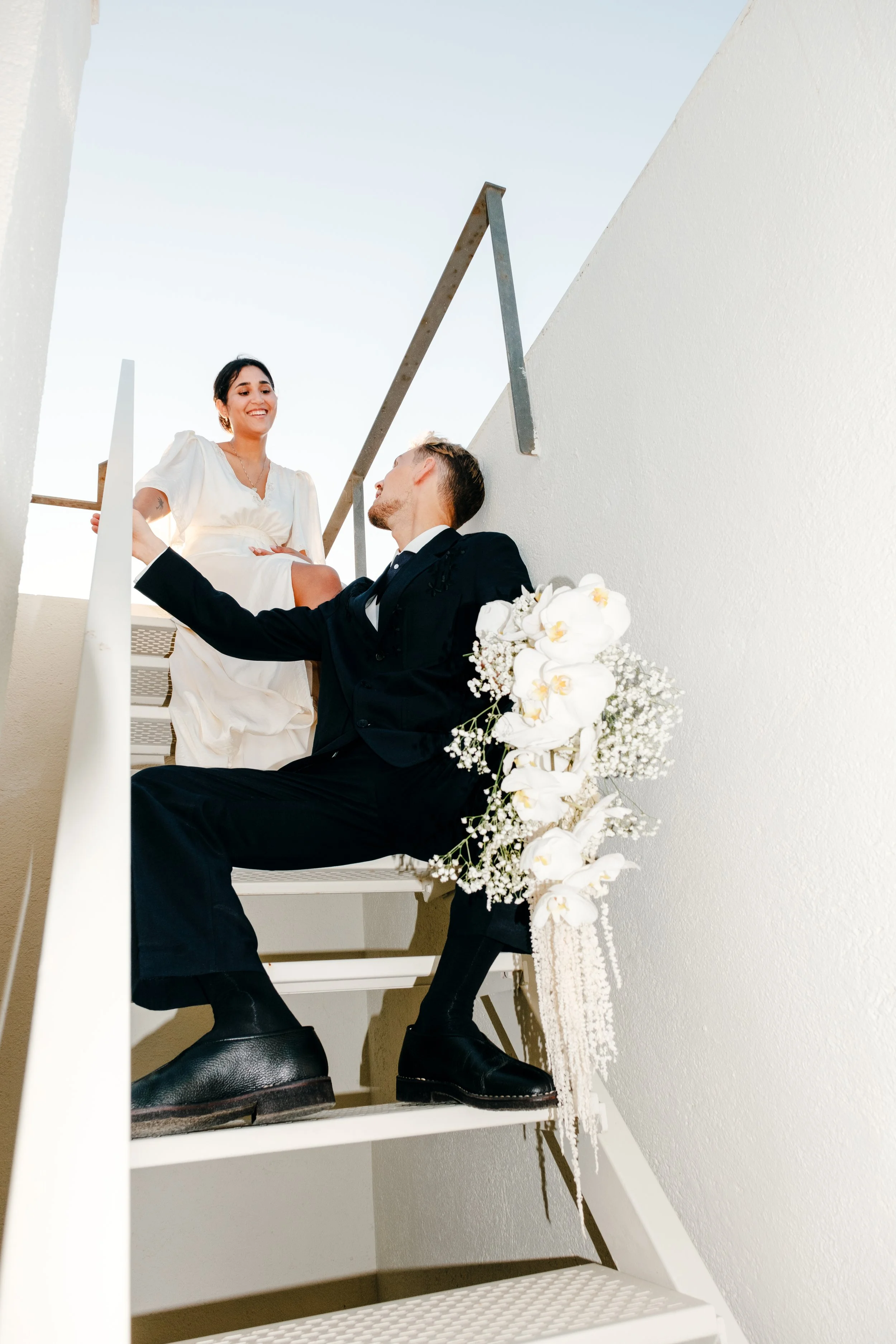 Ein Mann in formeller Kleidung und eine Frau in einem weißen Kleid sitzen und stehen auf einer Treppe, mit weißen Blumen als Dekoration, im Freien bei Tageslicht, in einer modernen Umgebung.