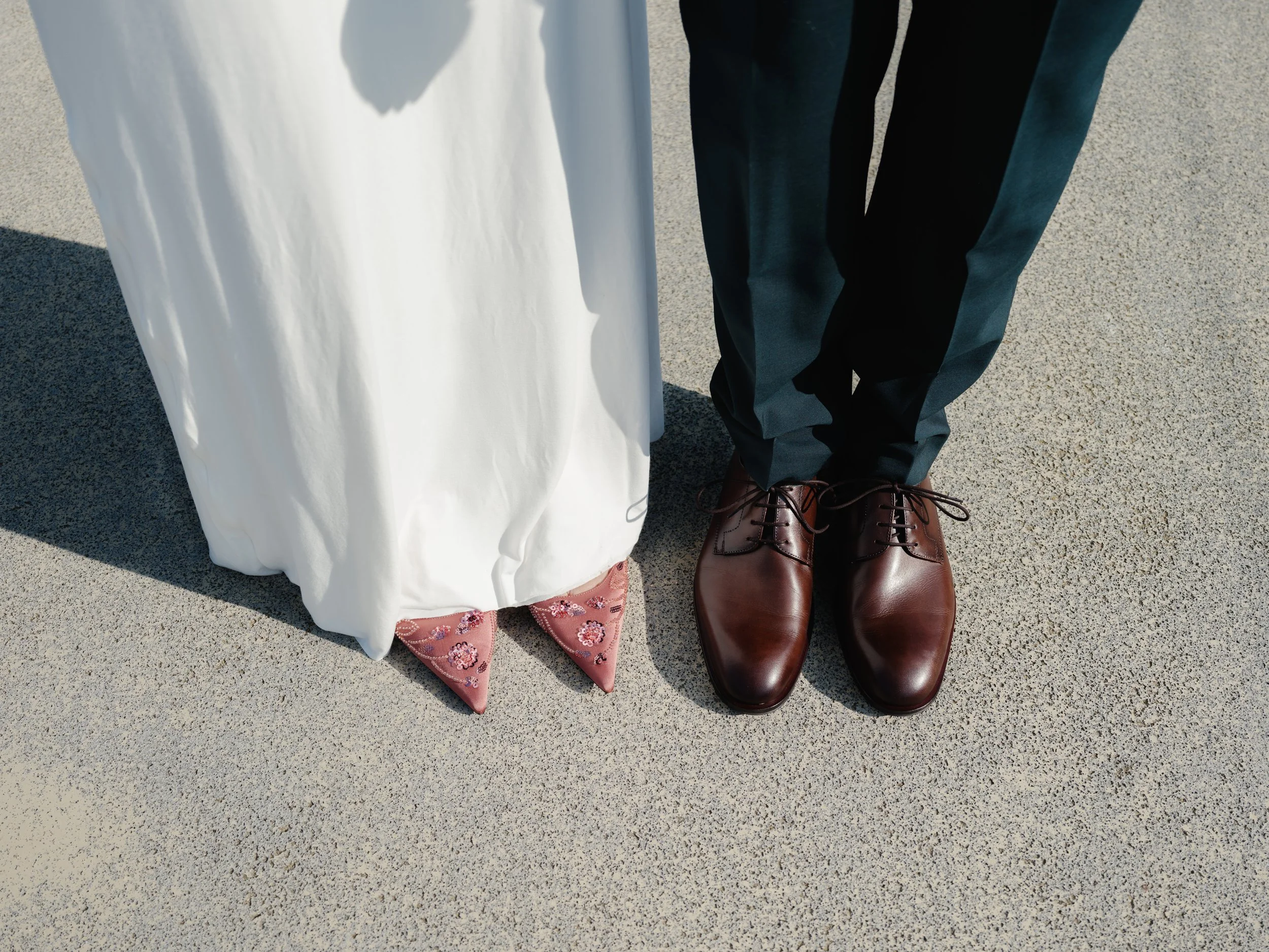 Nahaufnahme der unteren Körperteile eines Brautpaares, das auf einer betonierten Oberfläche steht, mit sichtbaren Hochzeitsschuhe und Hosen sowie einem Brautrock mit Stickerei.