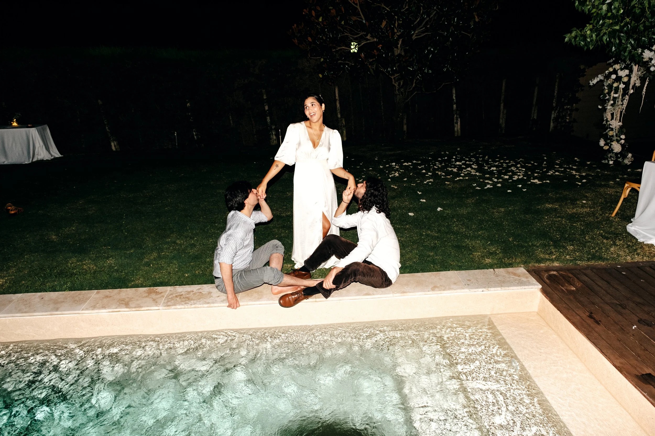 Menschen an einem Abend im Freien, zwei Männer sitzen auf dem Boden, eine Frau steht zwischen ihnen und hält ihre Hände, alle lächeln, hinter ihnen ist ein Garten mit Blättern und Dekorationen.