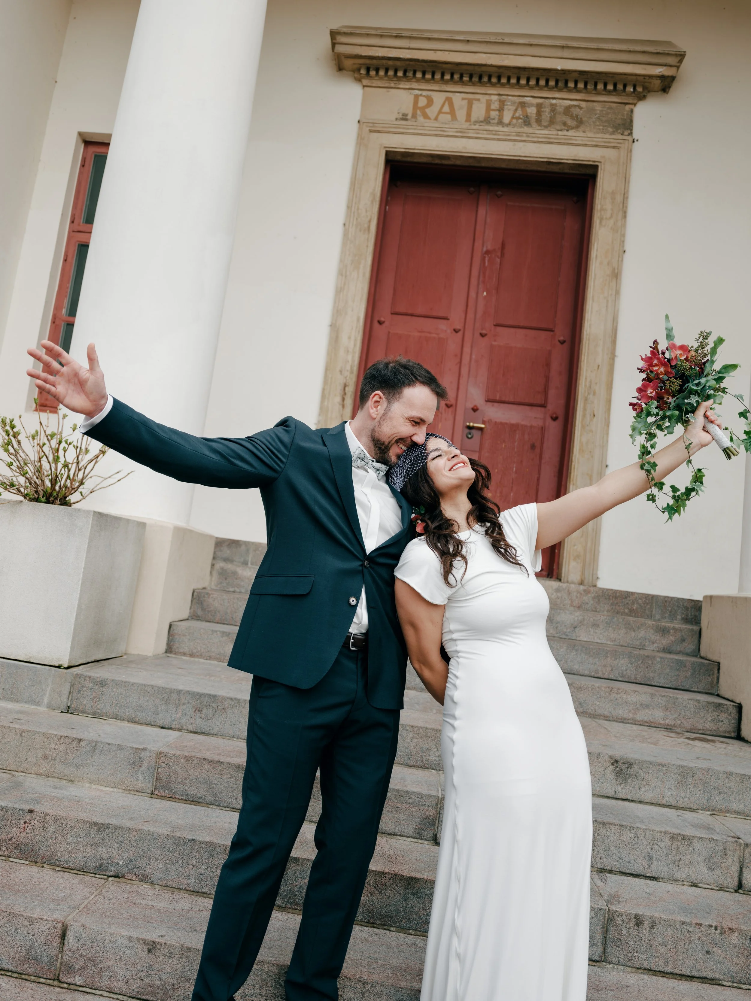 Ein verheiratetes Paar steht vor einem Rathaus, der Mann trägt einen dunklen Anzug, die Frau in einem weißen Kleid, sie hält einen Blumenstrauß hoch und sie lächeln glücklich.