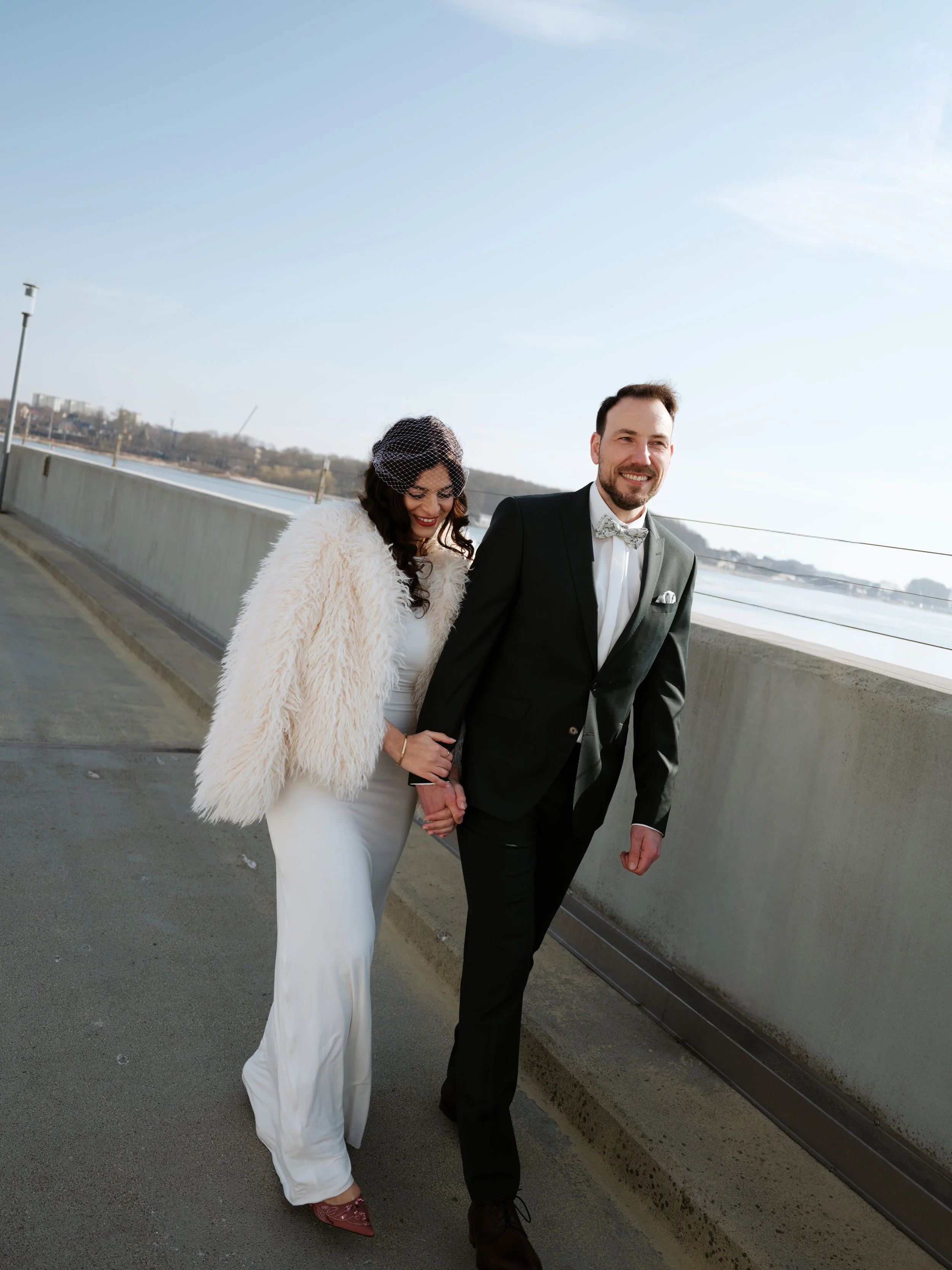 Ein Paar in formeller Kleidung, die Hand in Hand auf einer Brücke entlang spaziert, mit Fluss und Stadt im Hintergrund, sonniger Tag.
