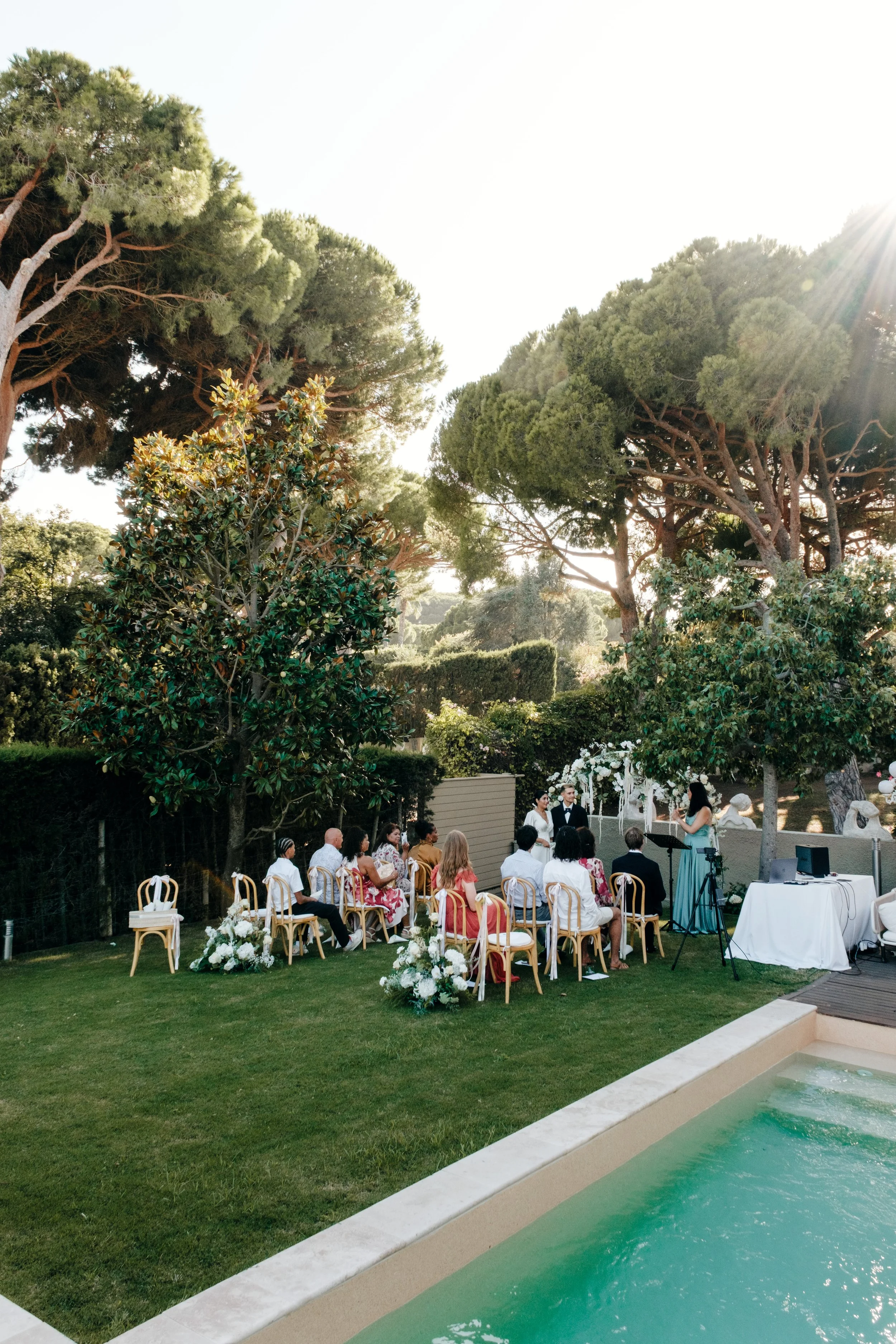 Eine Hochzeit im Freien mit Gästen, die auf Stühlen sitzen, während das Brautpaar vorne steht. Es gibt Blumen und Grün um sie herum, große Bäume im Hintergrund und das Sonnenlicht strahlt von rechts.