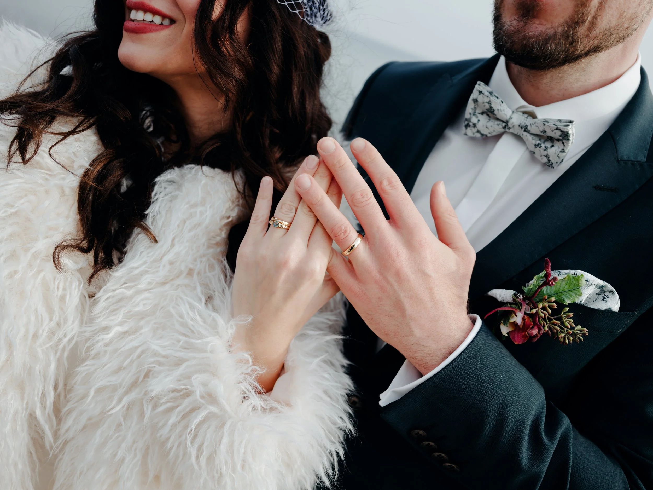 Ein frisch vermähltes Paar zeigt ihre Eheringe. Die Frau trägt einen weißen, pelzigen Mantel, der Mann trägt einen schwarzen Anzug mit Fliege und eine Blumenschleife.