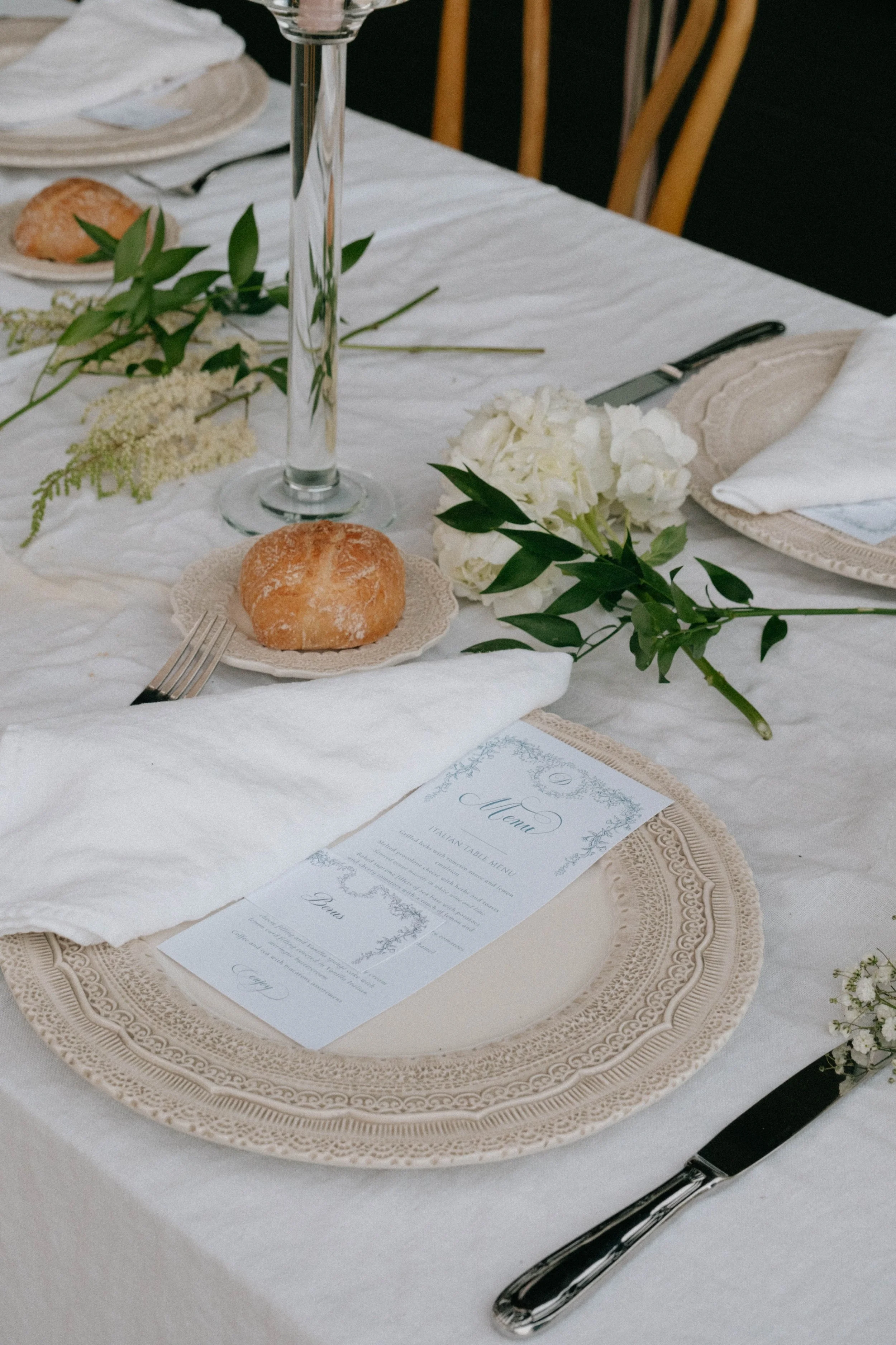 Eleganter gedeckter Tisch mit weißen Stofftischläufern, Tellern, Broten, Besteck, einer Kerze und Blumen, vermutlich für eine Feier oder Hochzeit.