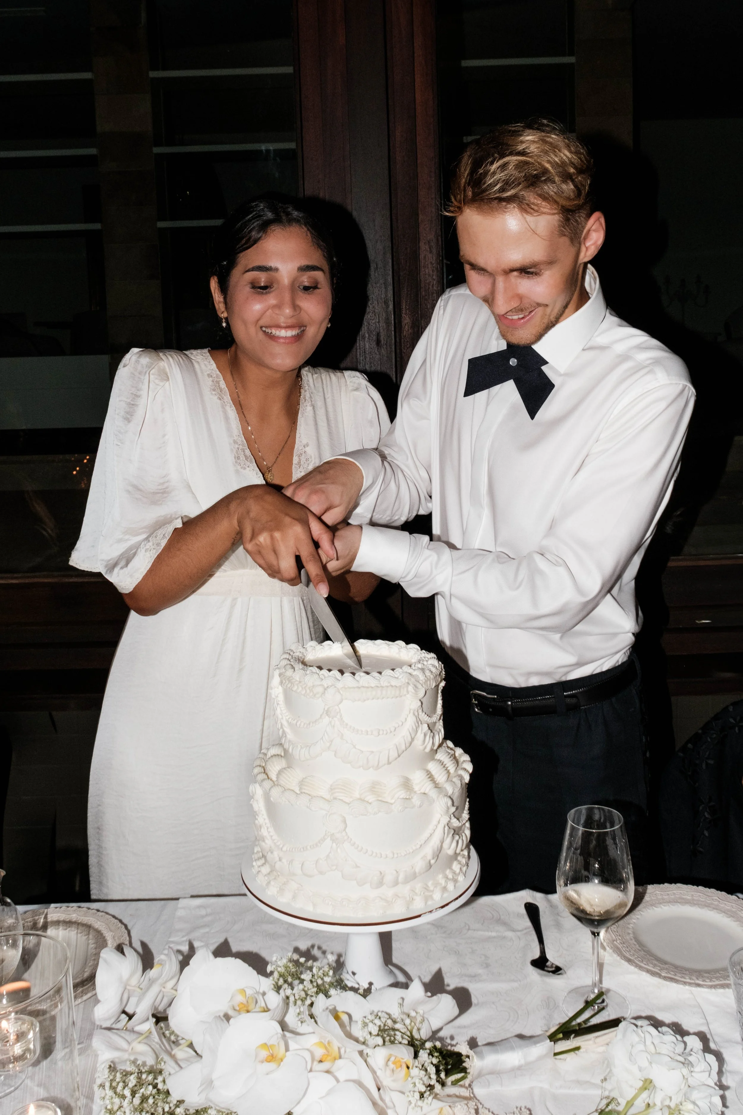 Ein Paar schneidet eine Hochzeitstorte bei einer Feier. Sie tragen formelle Kleidung, das Mädchen in einem weißen Kleid und der Junge in einem weißen Hemd mit Fliege.